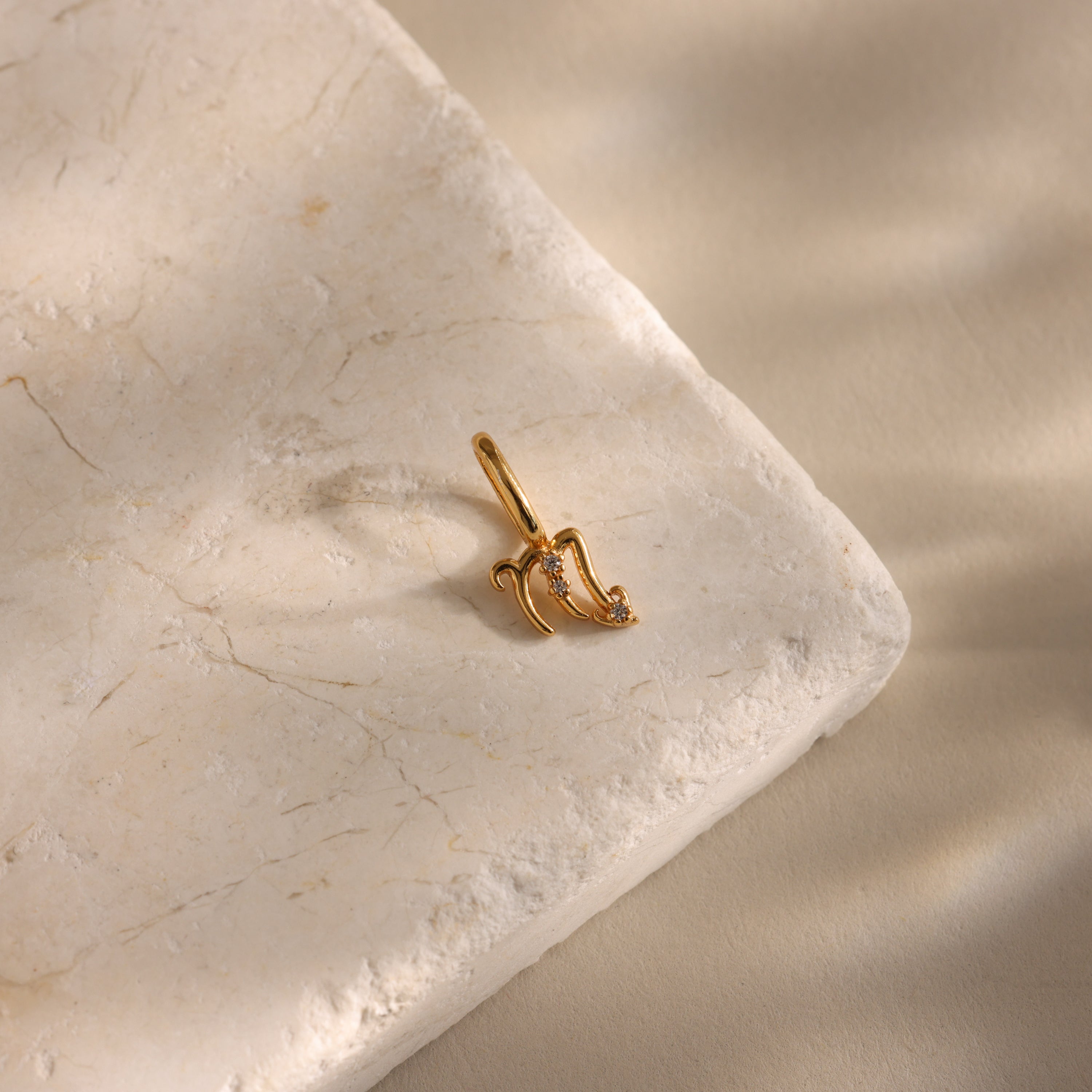 The Zodiac Charm pendant, adorned with small diamonds, rests on a textured beige stone and is softly lit—an elegant celestial jewelry piece.