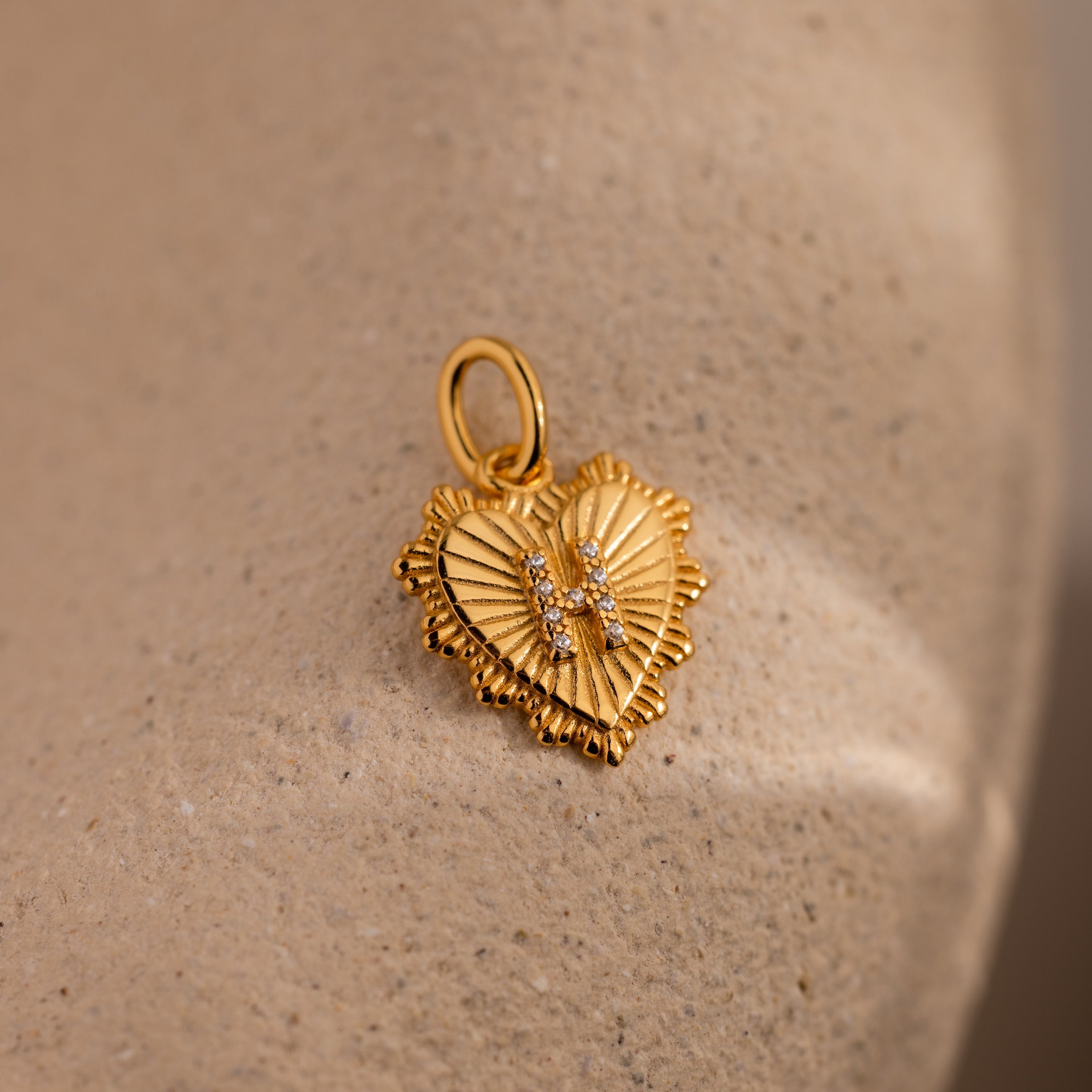The Ribbed Heart Pave Initial Charm features textured edges and sparkling gemstones, ideal for personalized jewelry lovers, shown on a beige, textured background.