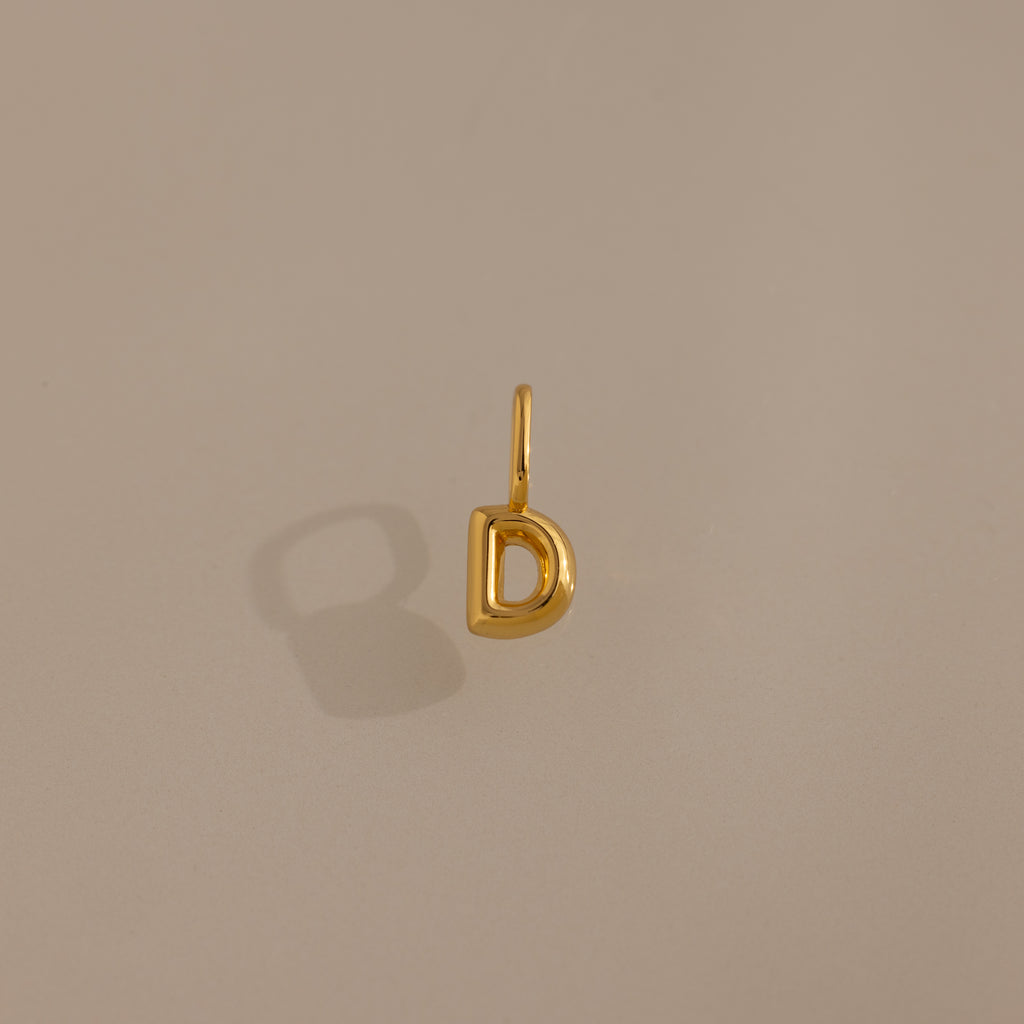 Bubble Letter Charm in 18K Gold D pendant displayed on a neutral beige background with a soft left shadow—ideal personalized jewelry or a charming addition for bracelet enthusiasts.