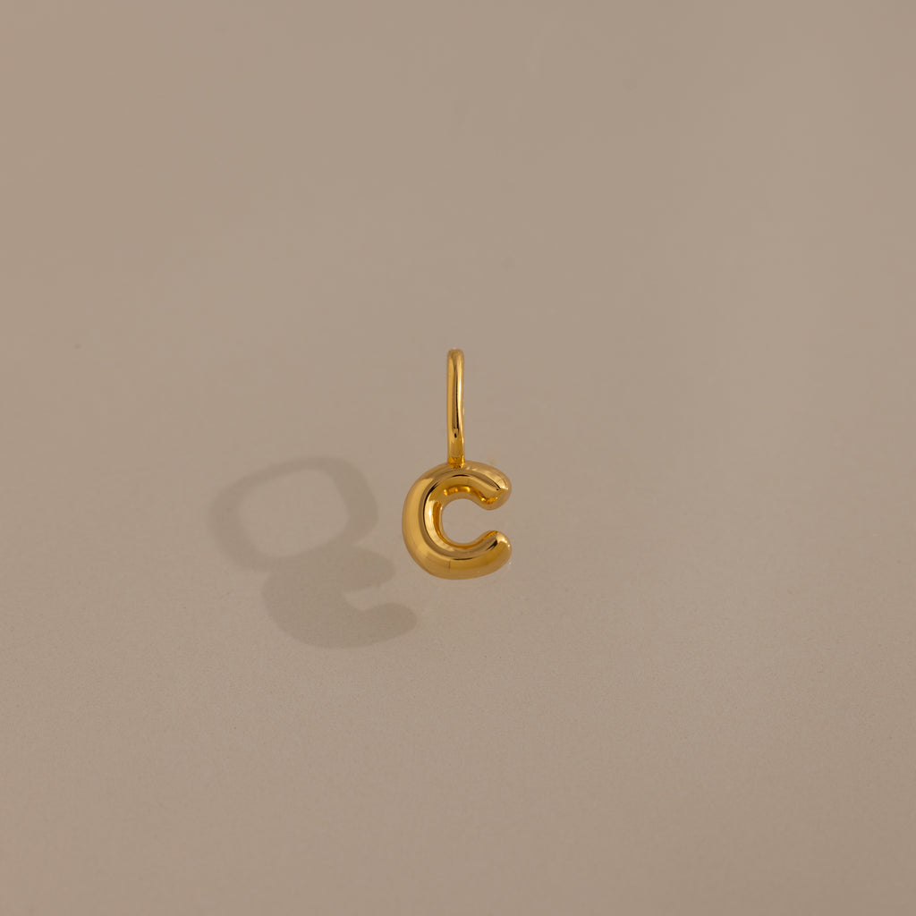The Bubble Letter Charm in 18K Gold shaped like the letter C casts a shadow on a beige background, making it an ideal personalized jewelry piece or bracelet charm.