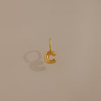 The Bubble Letter Charm in 18K Gold shaped like the letter C casts a shadow on a beige background, making it an ideal personalized jewelry piece or bracelet charm.
