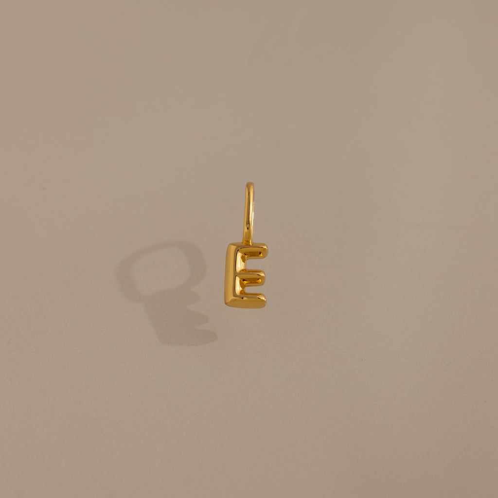 The Bubble Letter Charm in 18K Gold, shaped like the letter E, casts a shadow on a plain beige background—an elegant piece of personalized jewelry.