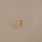 The Bubble Letter Charm in 18K Gold, shaped like the letter E, casts a shadow on a plain beige background—an elegant piece of personalized jewelry.