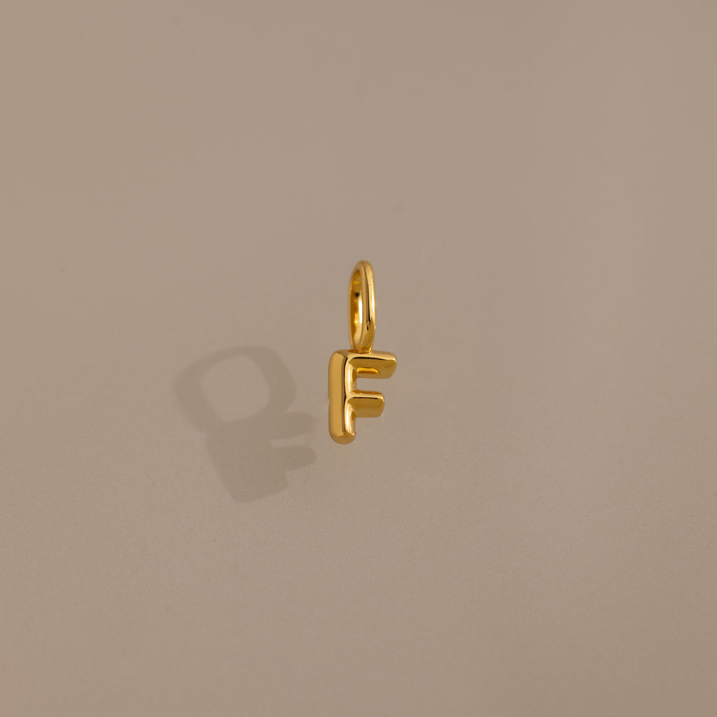 The Bubble Letter Charm in 18K Gold, shaped like the letter F, sits on a beige background—ideal as personalized jewelry or a distinctive charm for bracelets.