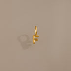 The Bubble Letter Charm in 18K Gold, shaped like the letter F, sits on a beige background—ideal as personalized jewelry or a distinctive charm for bracelets.