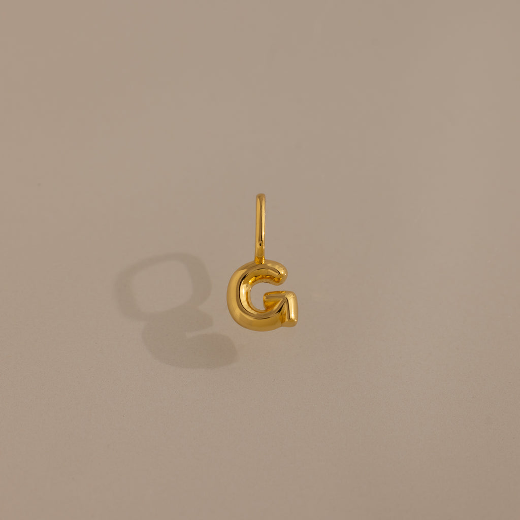 The Bubble Letter Charm in 18K Gold, shaped as the letter G, offers a stylish personalized jewelry option and is showcased on a simple beige background.