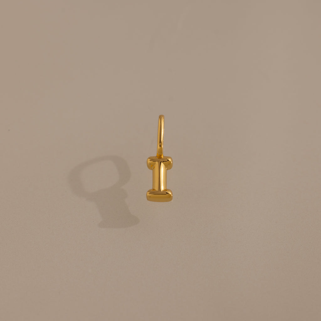 The Bubble Letter Charm in 18K Gold, shaped like the letter I, rests on a plain beige background—an elegant piece of personalized jewelry.