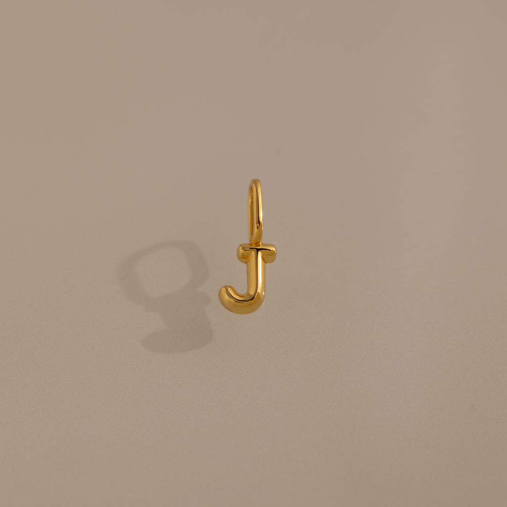 The Bubble Letter Charm in 18K Gold, shaped like the letter J and photographed on a plain beige background, makes a perfect personalized jewelry piece or a charming addition to your bracelet.