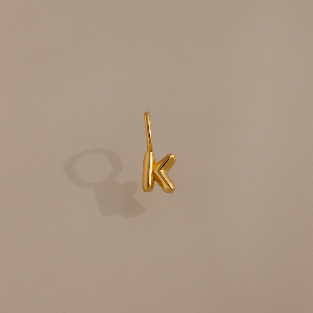 The Bubble Letter Charm in 18K Gold, shaped as a K, makes a stylish personalized pendant or bracelet charm and is showcased on a neutral beige background with a soft shadow.