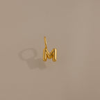 Bubble Letter Charm in 18K Gold—a gold bubble letter M charm for bracelets, displayed on a beige background with a soft left-side shadow. Ideal for adding personalized style to your jewelry collection.