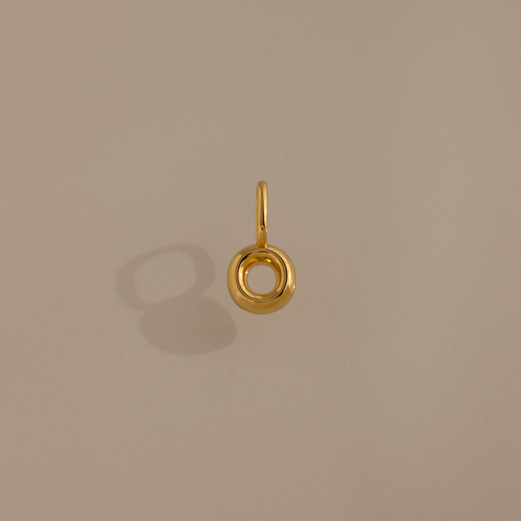 The Bubble Letter Charm in 18K Gold features a gold circular design with a small loop, shown casting a shadow on a beige background—ideal as a bracelet charm or for personalized jewelry.