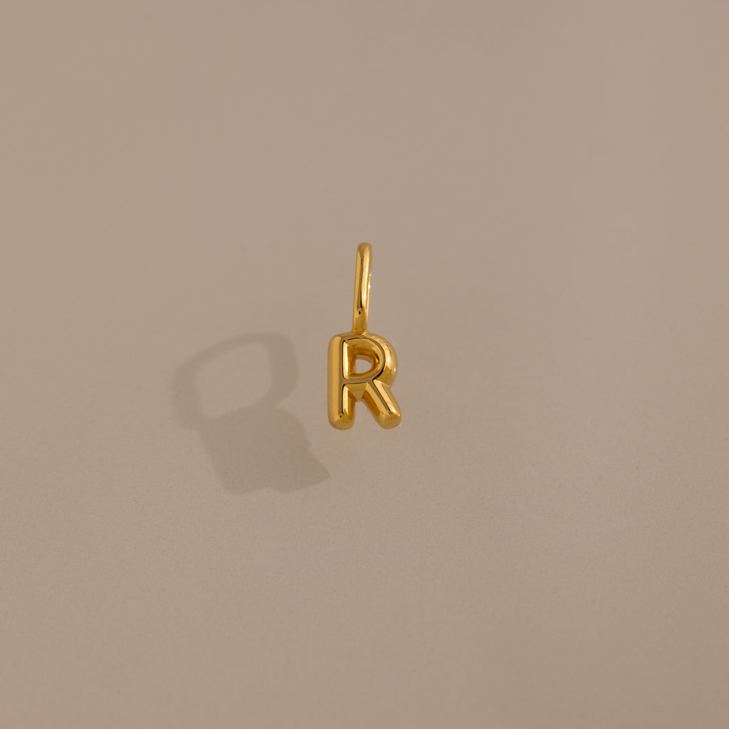 The Bubble Letter Charm in 18K Gold is a playful R-shaped pendant on a plain beige background with a subtle shadow—ideal as personalized jewelry or a unique charm for bracelet lovers.