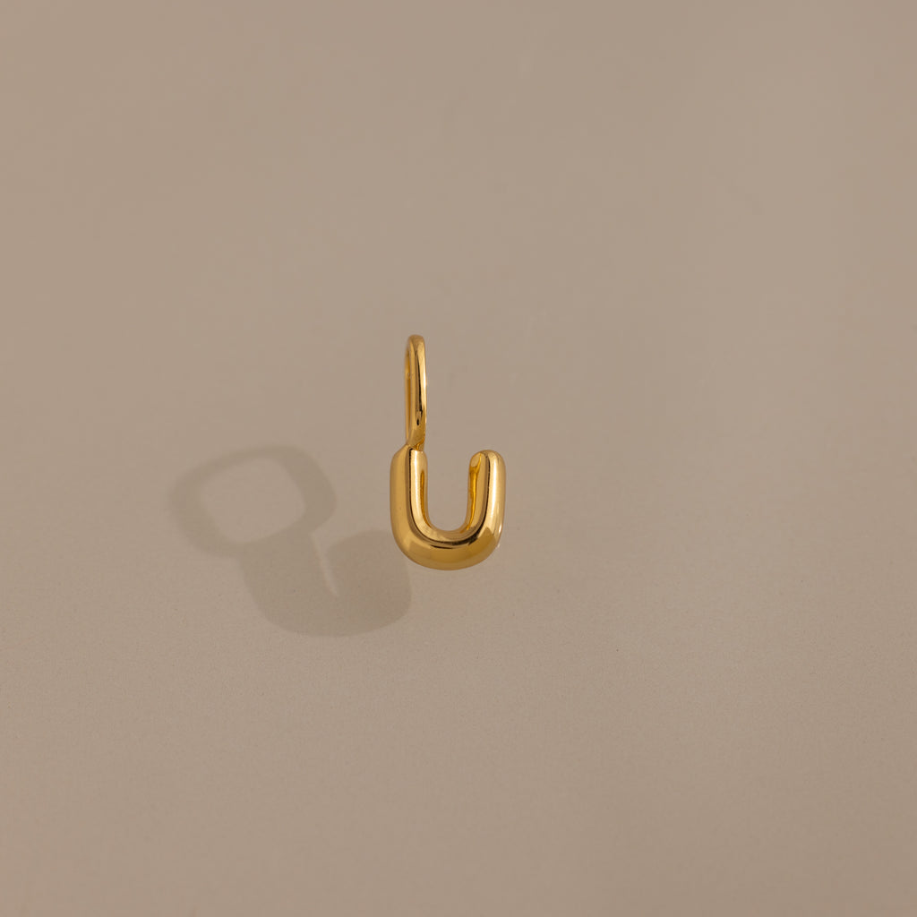 The Bubble Letter Charm in 18K Gold forms a striking U-shape on a beige surface, making it an ideal personalized jewelry piece or charm for bracelet lovers.