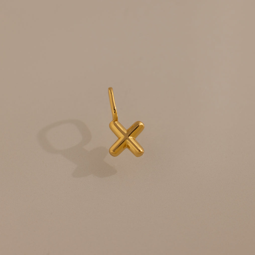 The Bubble Letter Charm in 18K Gold rests on a beige background, casting a shadow left—ideal for personalized jewelry or as a unique addition to any bracelet collection.