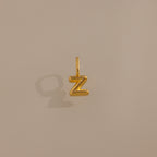 The Bubble Letter Charm in 18K Gold, shaped like a Z and casting a shadow on a beige surface, makes for a standout piece of personalized jewelry.