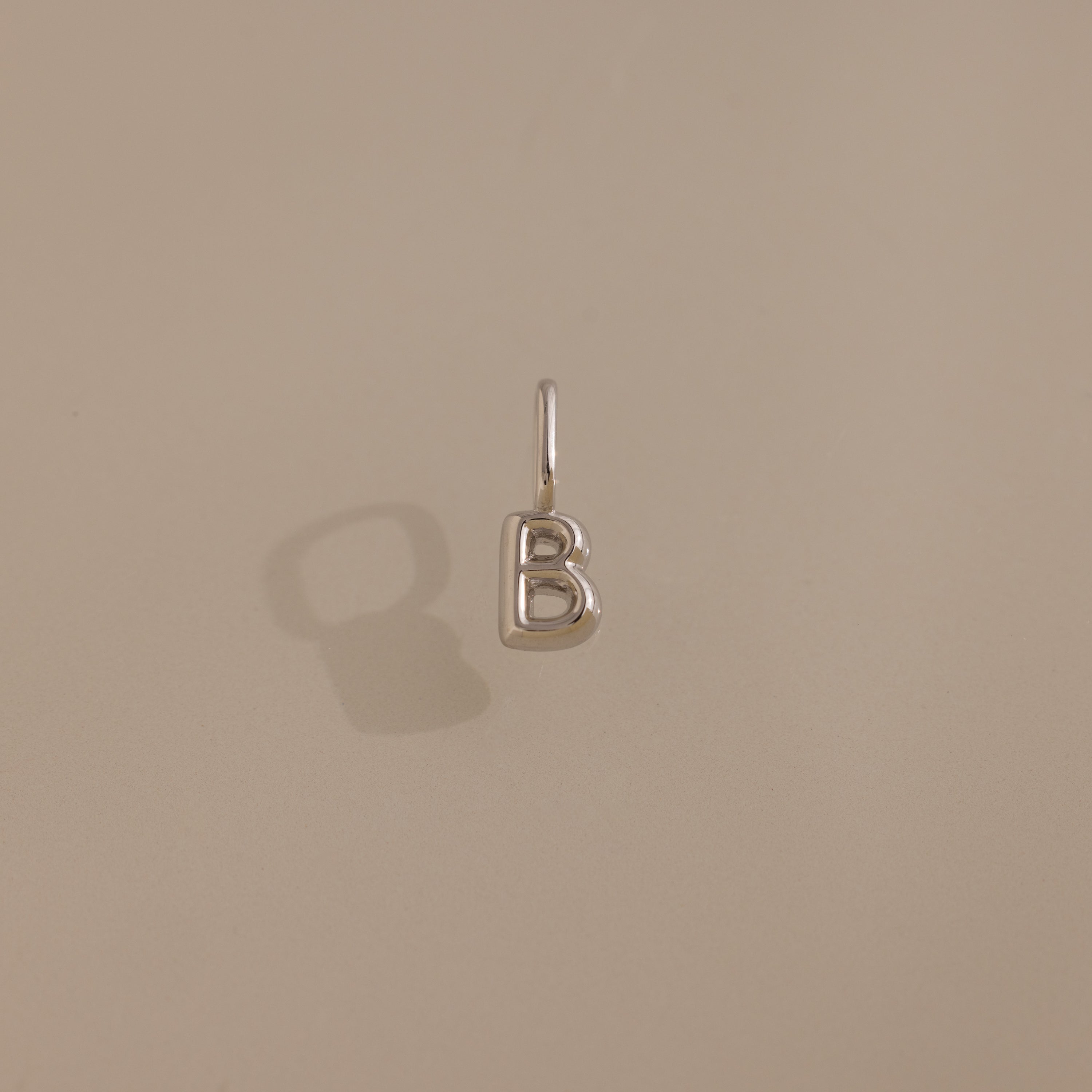 The Bubble Letter Charm in Sterling Silver, shaped like the letter B, rests on a beige surface—ideal for anyone who loves personalized jewelry.
