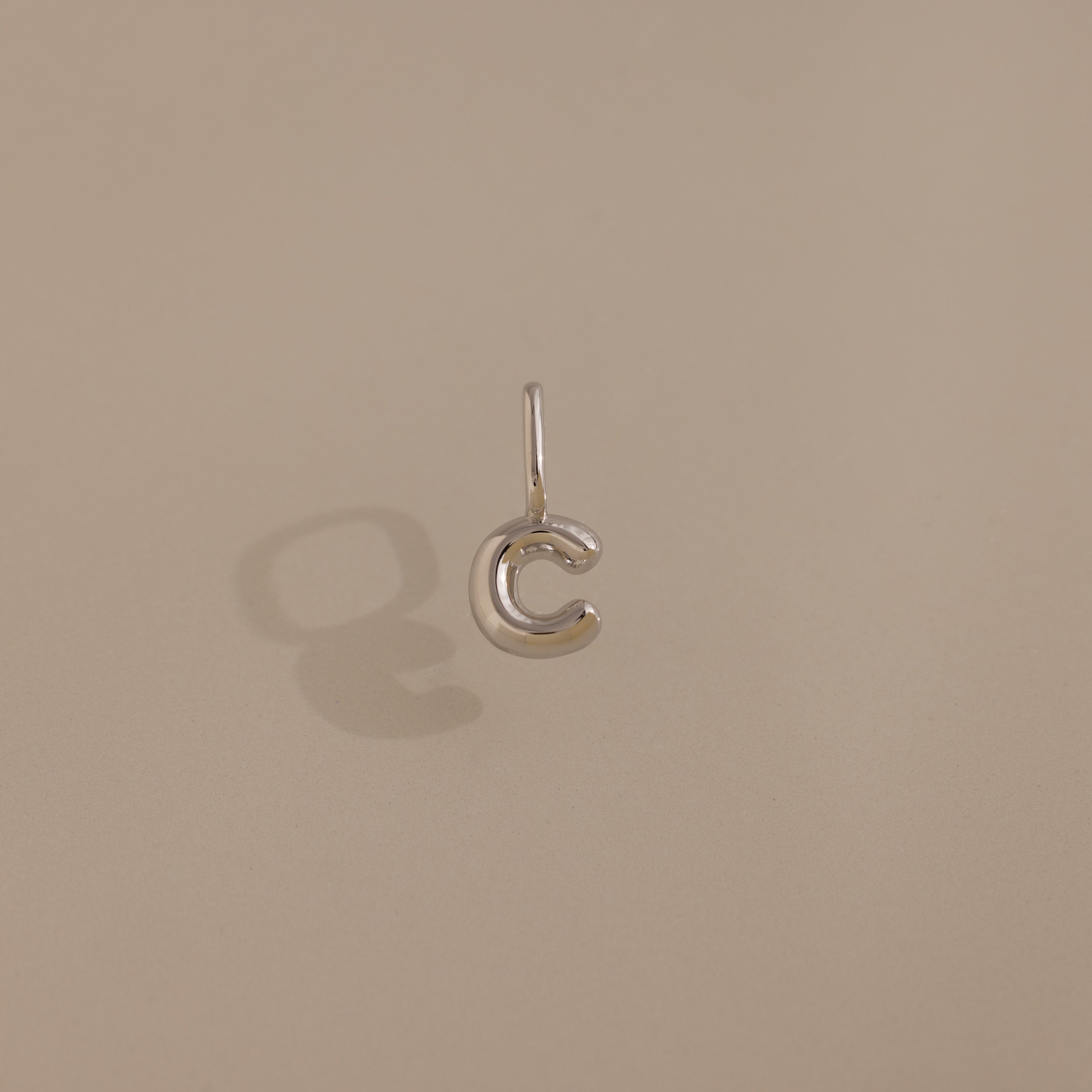 The Bubble Letter Charm in Sterling Silver, shaped like the letter “C,” casts a shadow on a beige background—adding a stylish, personalized touch to your jewelry collection.