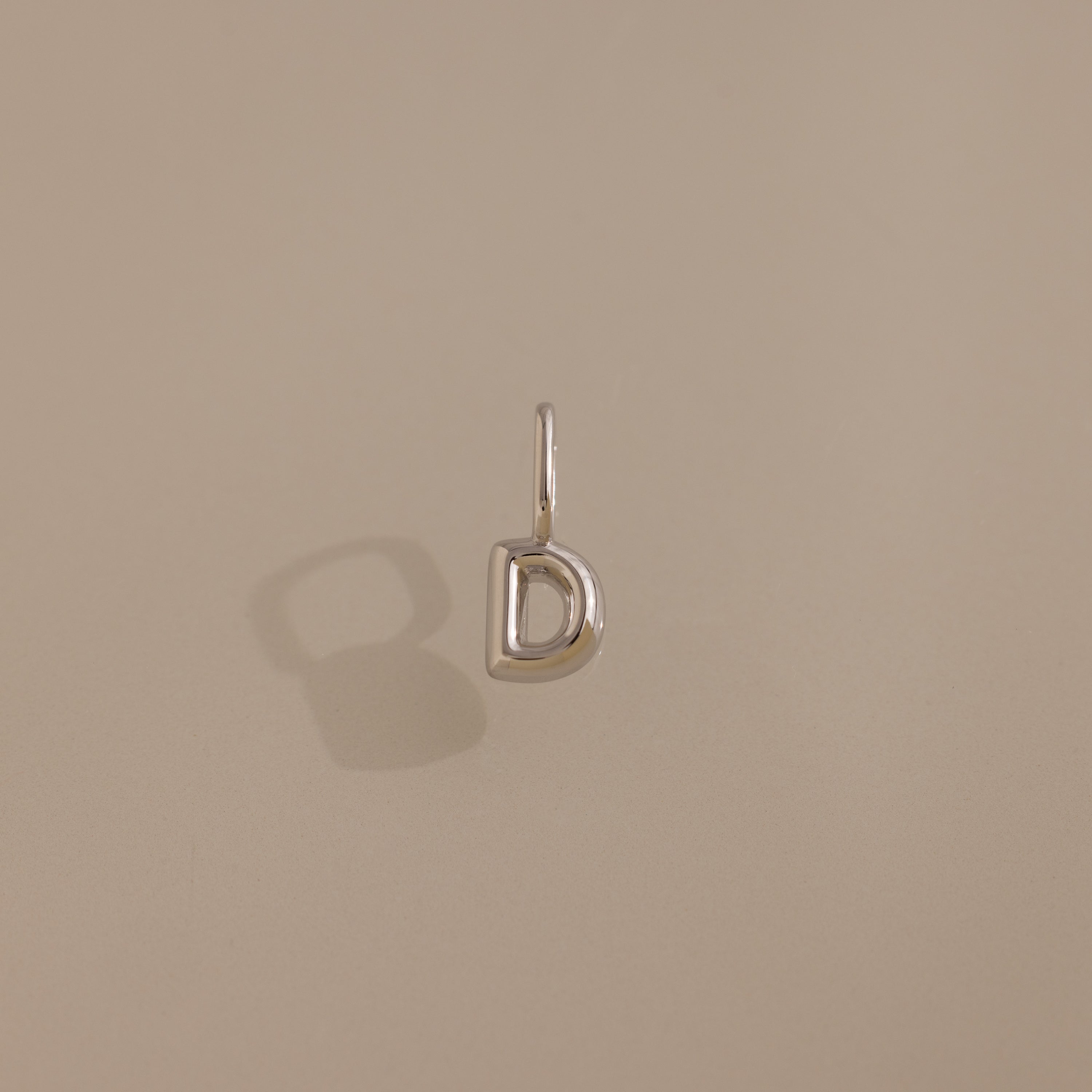 The Bubble Letter Charm in Sterling Silver, shaped like the letter D, is displayed on a plain beige background.