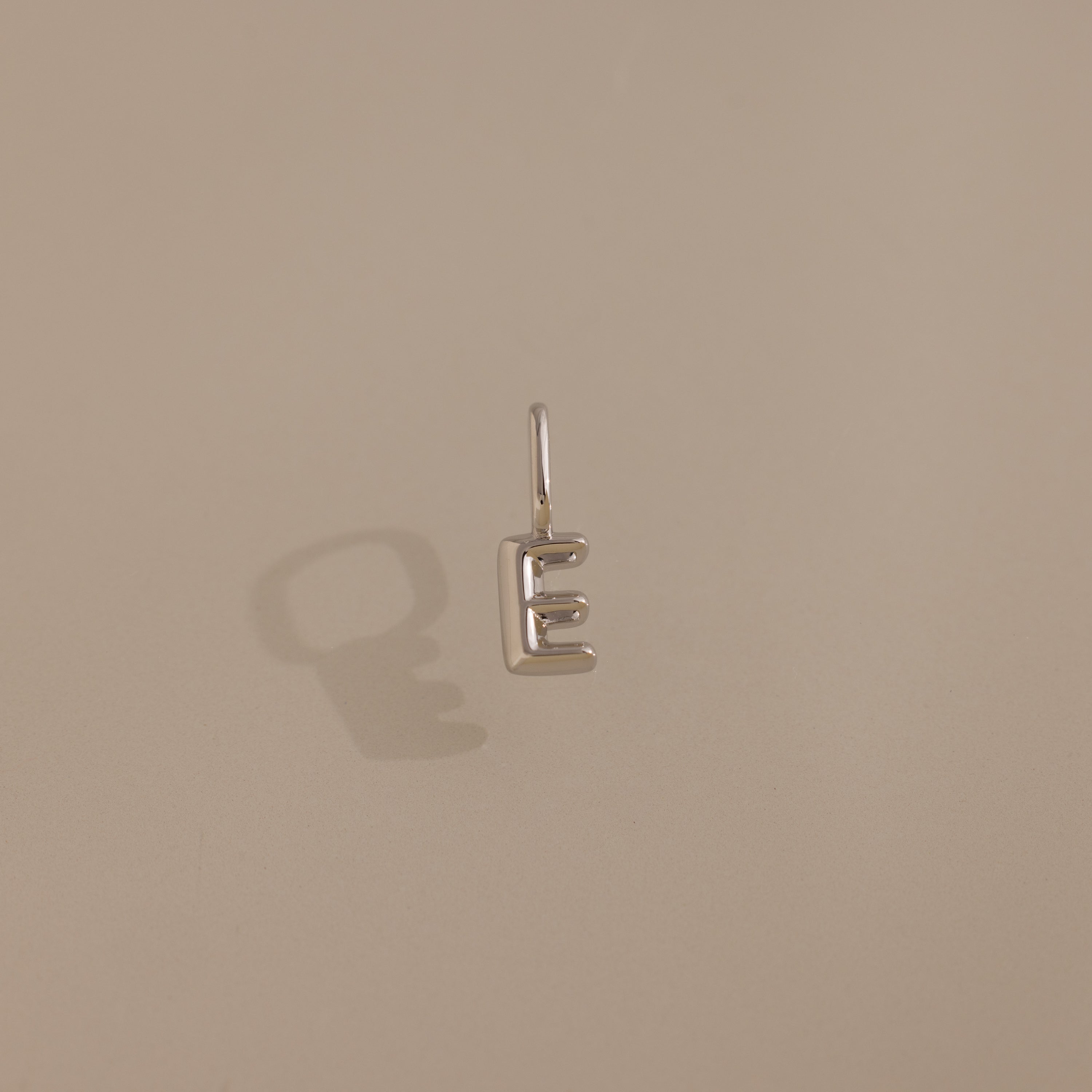 The Bubble Letter Charm in Sterling Silver, shaped like the letter E, casts a shadow on a beige surface.