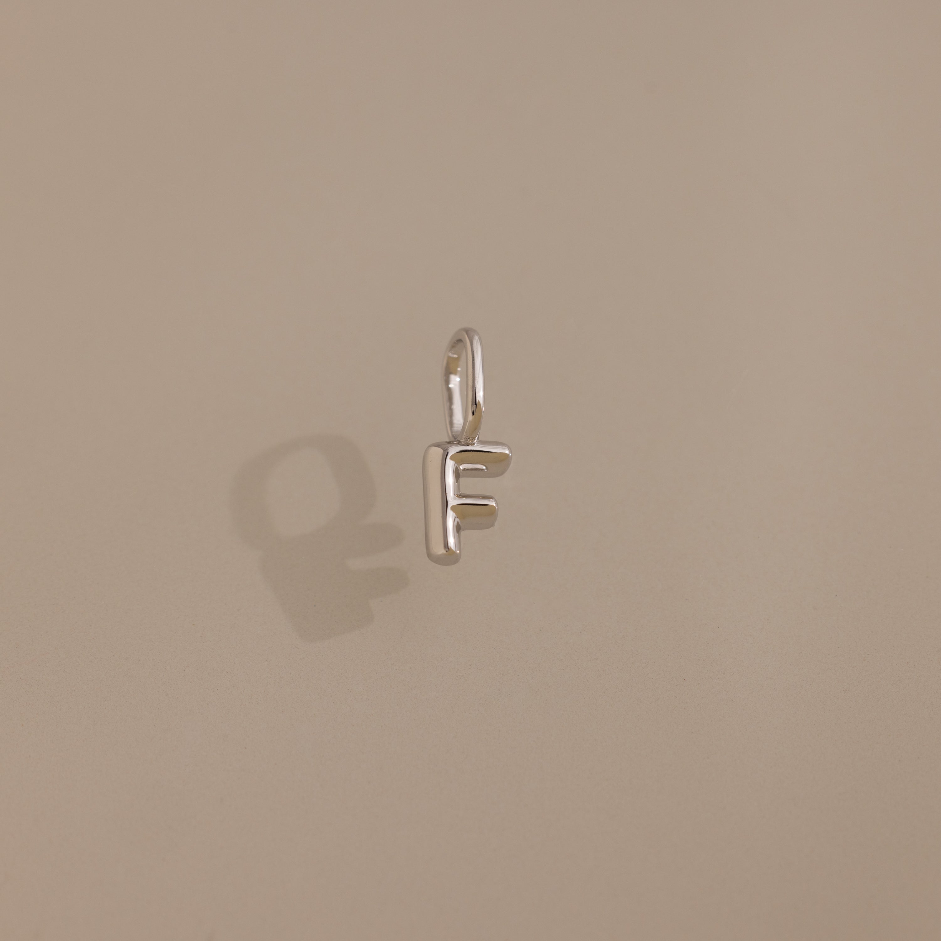 A sterling silver Bubble Letter Charm shaped like the letter F casts a shadow on a beige surface, capturing playful elegance for personalized jewelry lovers.
