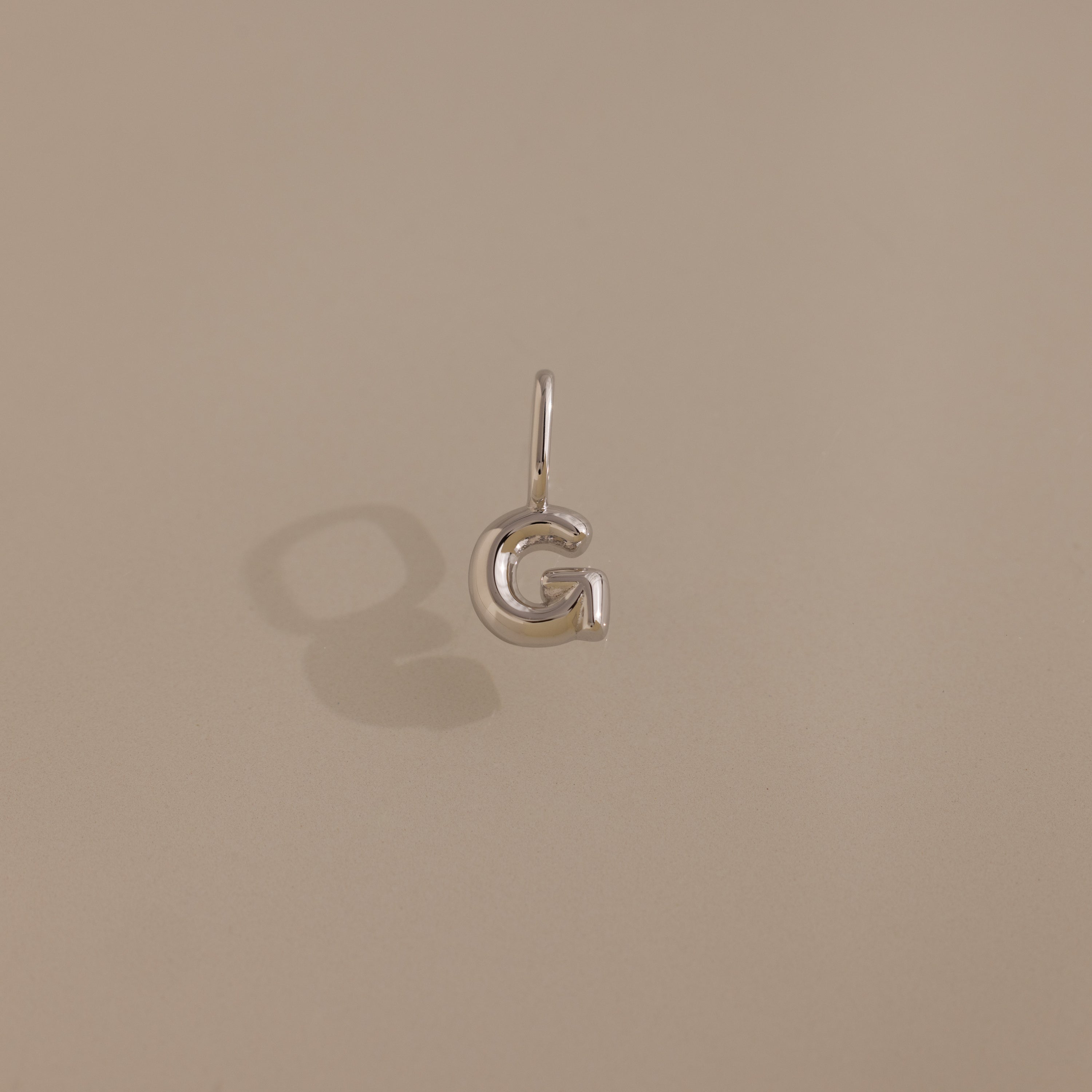 The Bubble Letter Charm in Sterling Silver, shaped like the letter G and shown on a beige background, is ideal for anyone seeking stylish personalized jewelry.