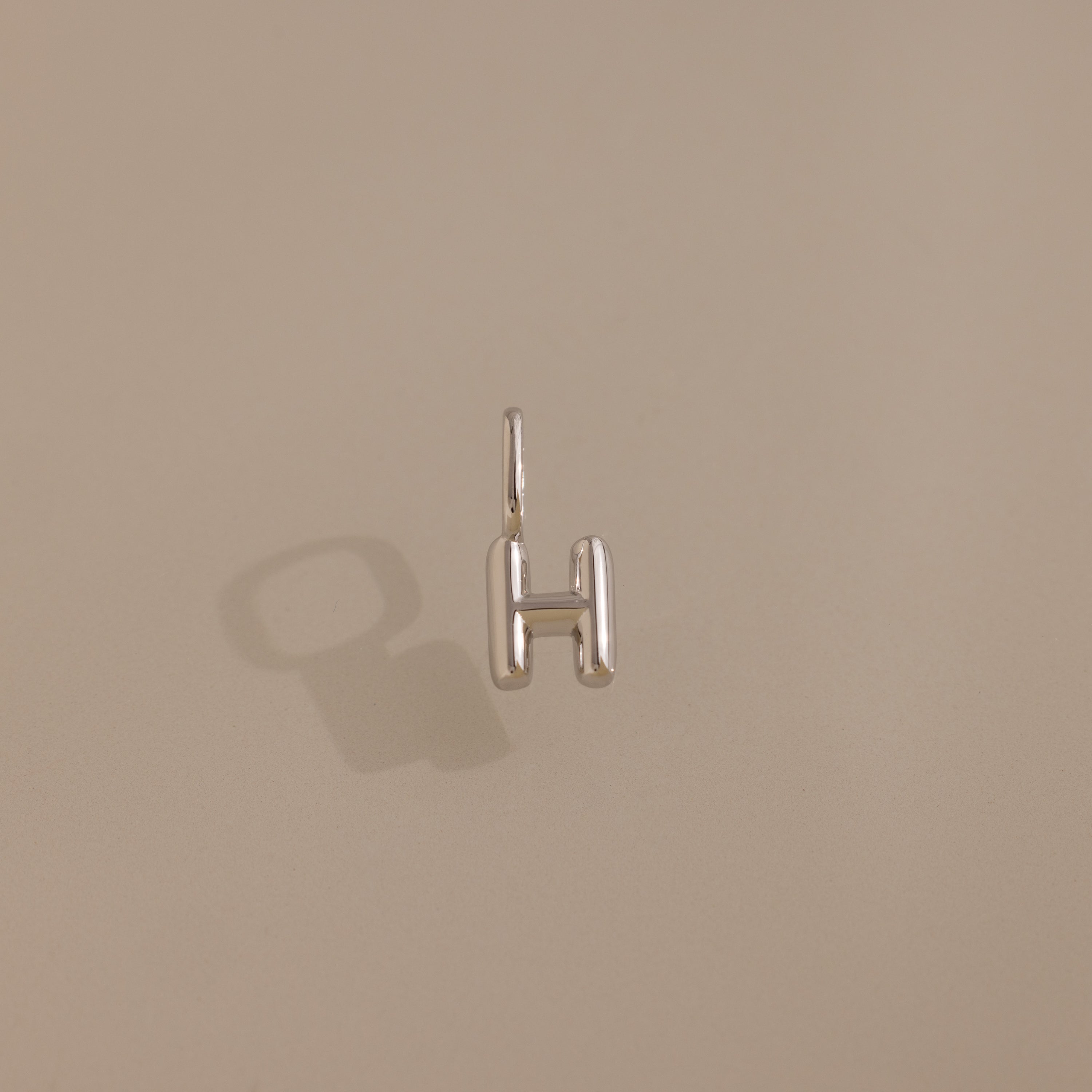 The Bubble Letter Charm in Sterling Silver, shaped like an "H," is displayed on a plain beige background with a visible shadow—ideal for fans of personalized jewelry.