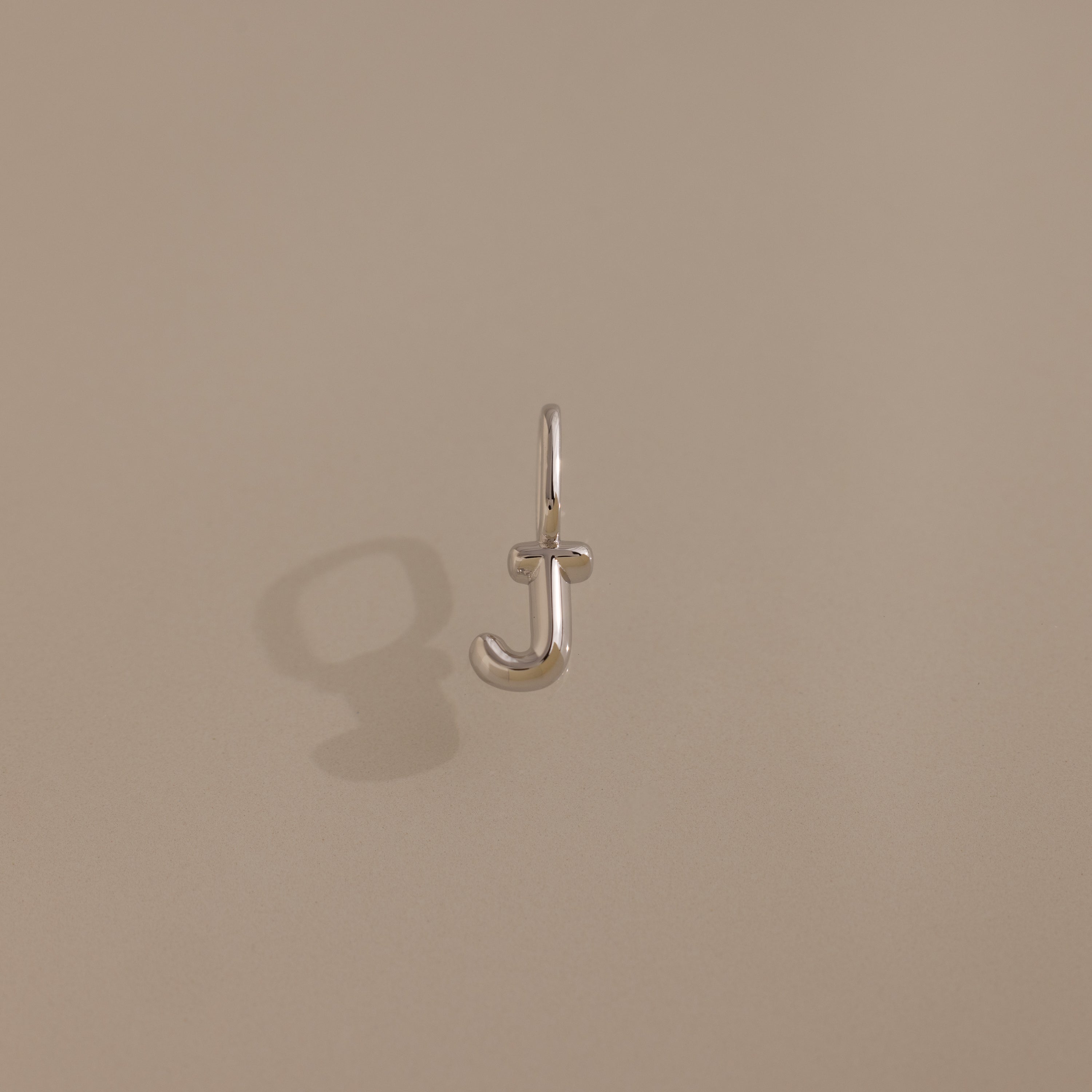 The Bubble Letter Charm in Sterling Silver features a "J" pendant casting a shadow on a plain beige background—a sleek, personalized jewelry piece.