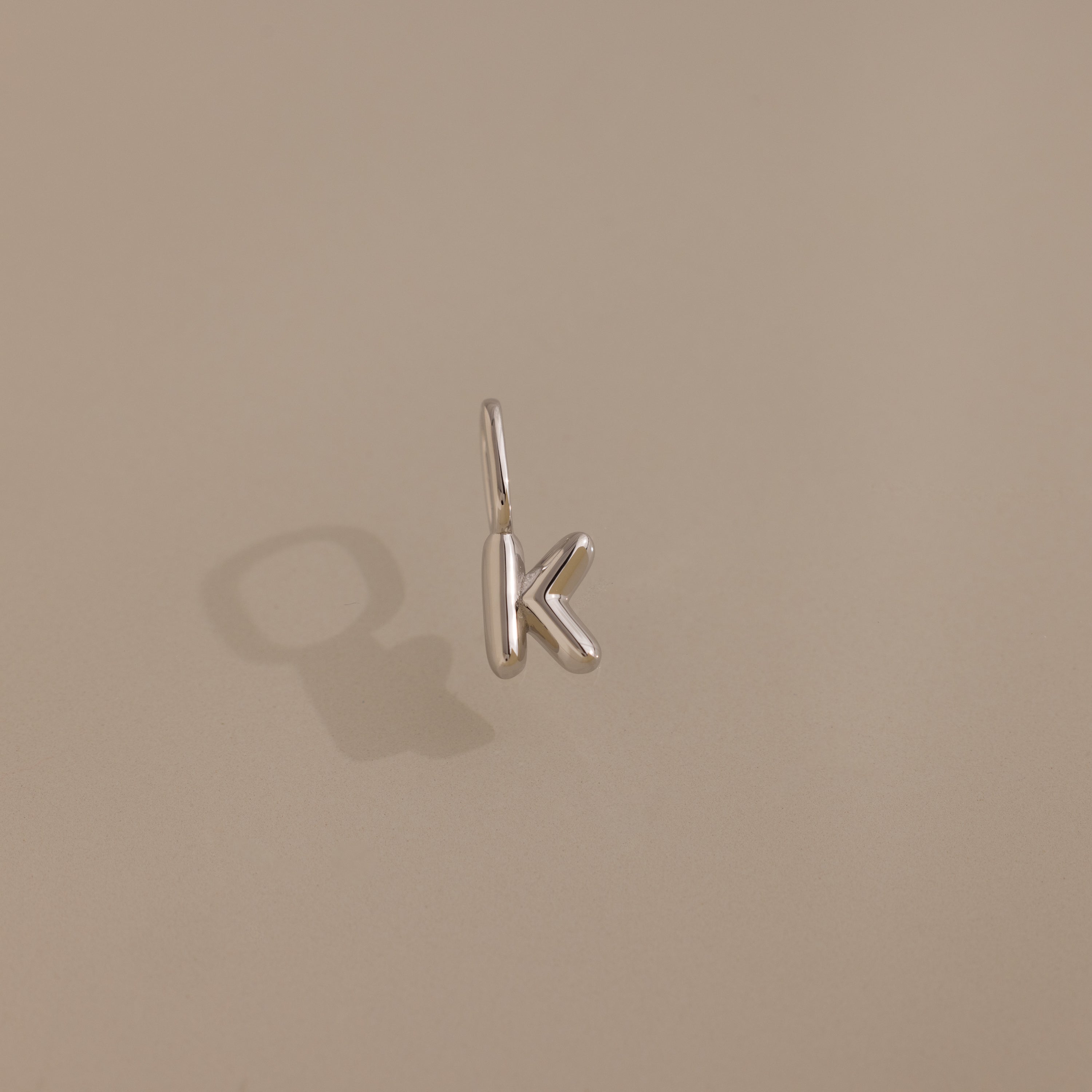The Bubble Letter Charm in Sterling Silver, shaped as a bold "K," shines on a beige background—ideal for anyone who loves personalized jewelry.