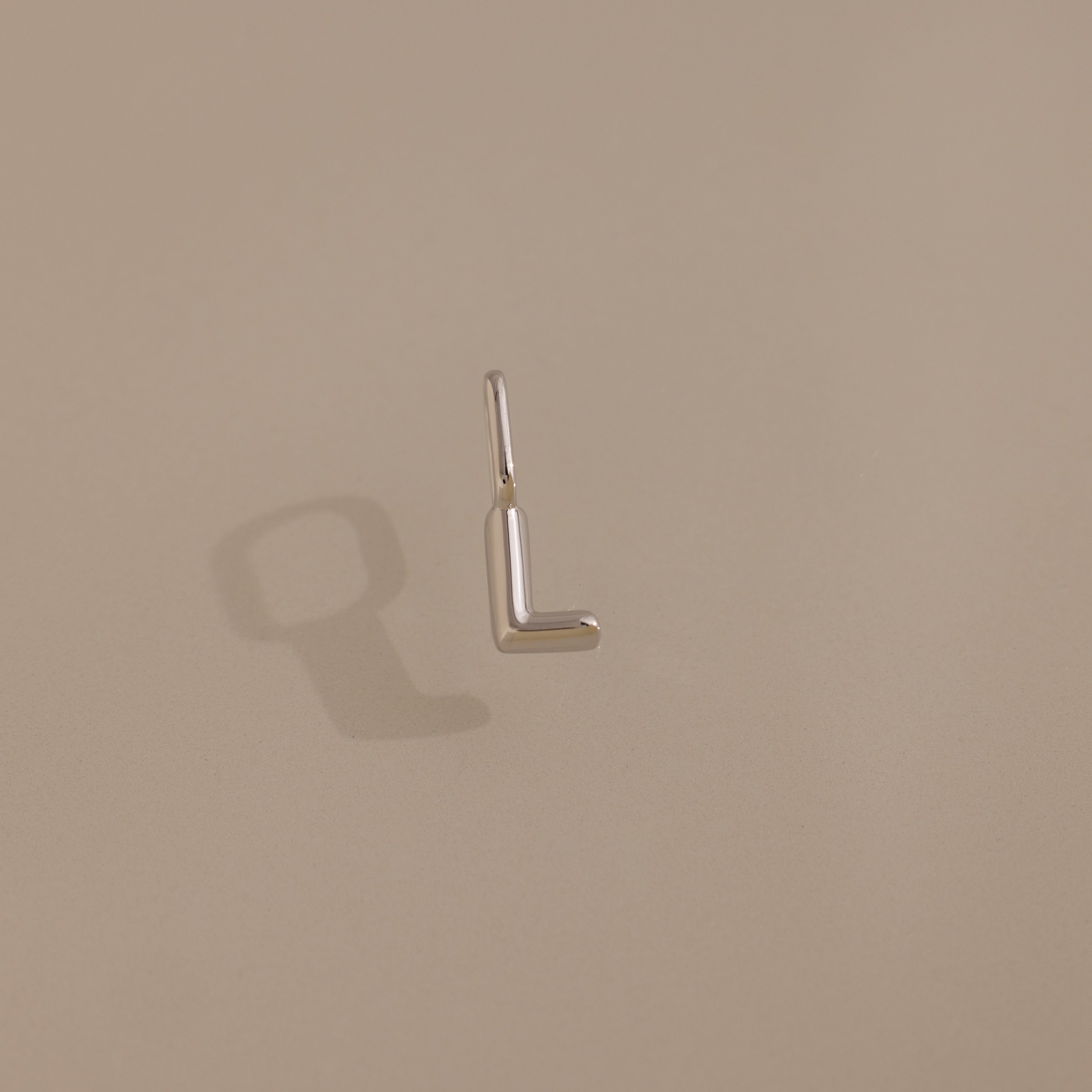 The Bubble Letter Charm in Sterling Silver, shaped like the letter L, rests on a beige background with its shadow visible.