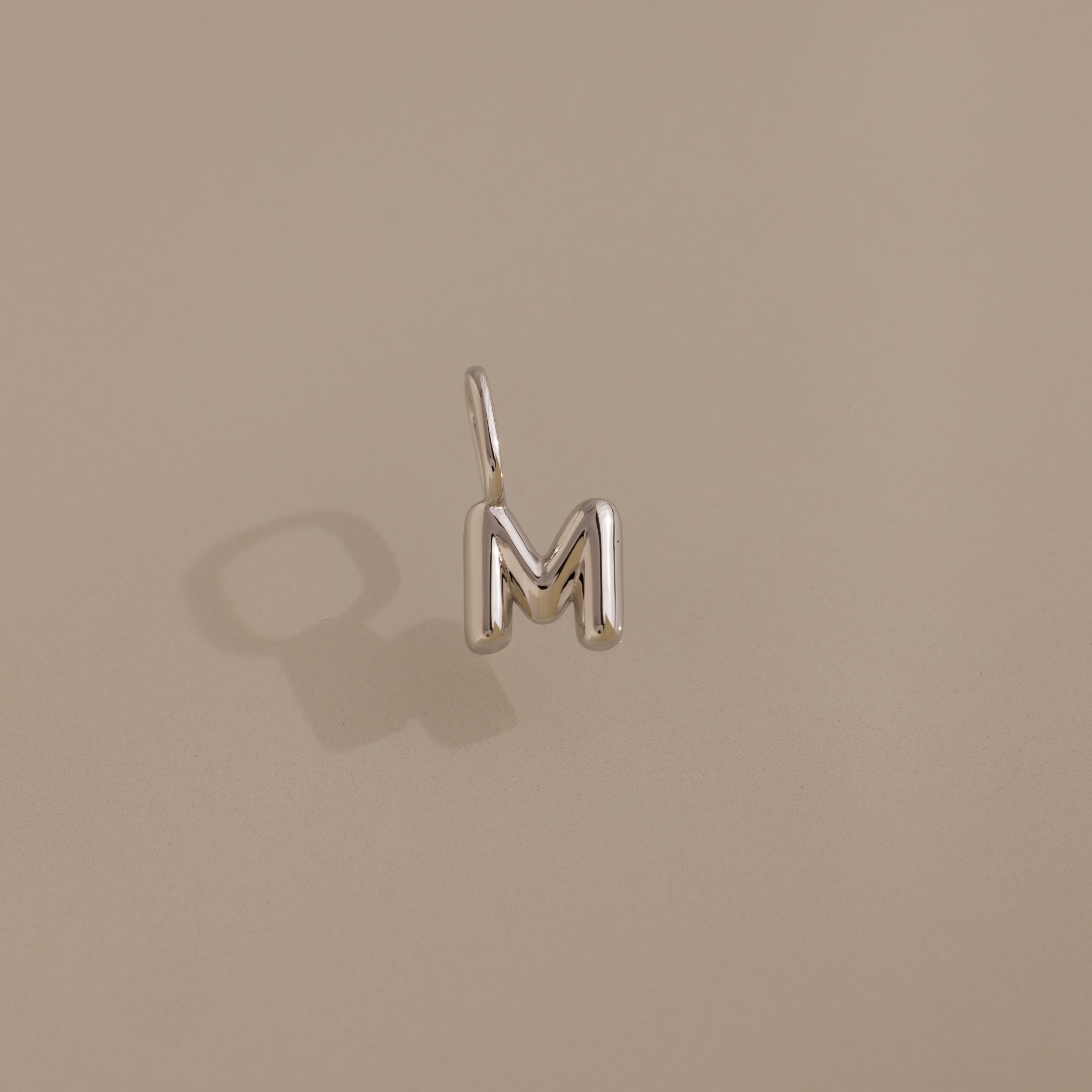 The Bubble Letter Charm in Sterling Silver, shaped like the letter M, rests on a beige surface and casts a soft shadow—an ideal personalized jewelry piece.