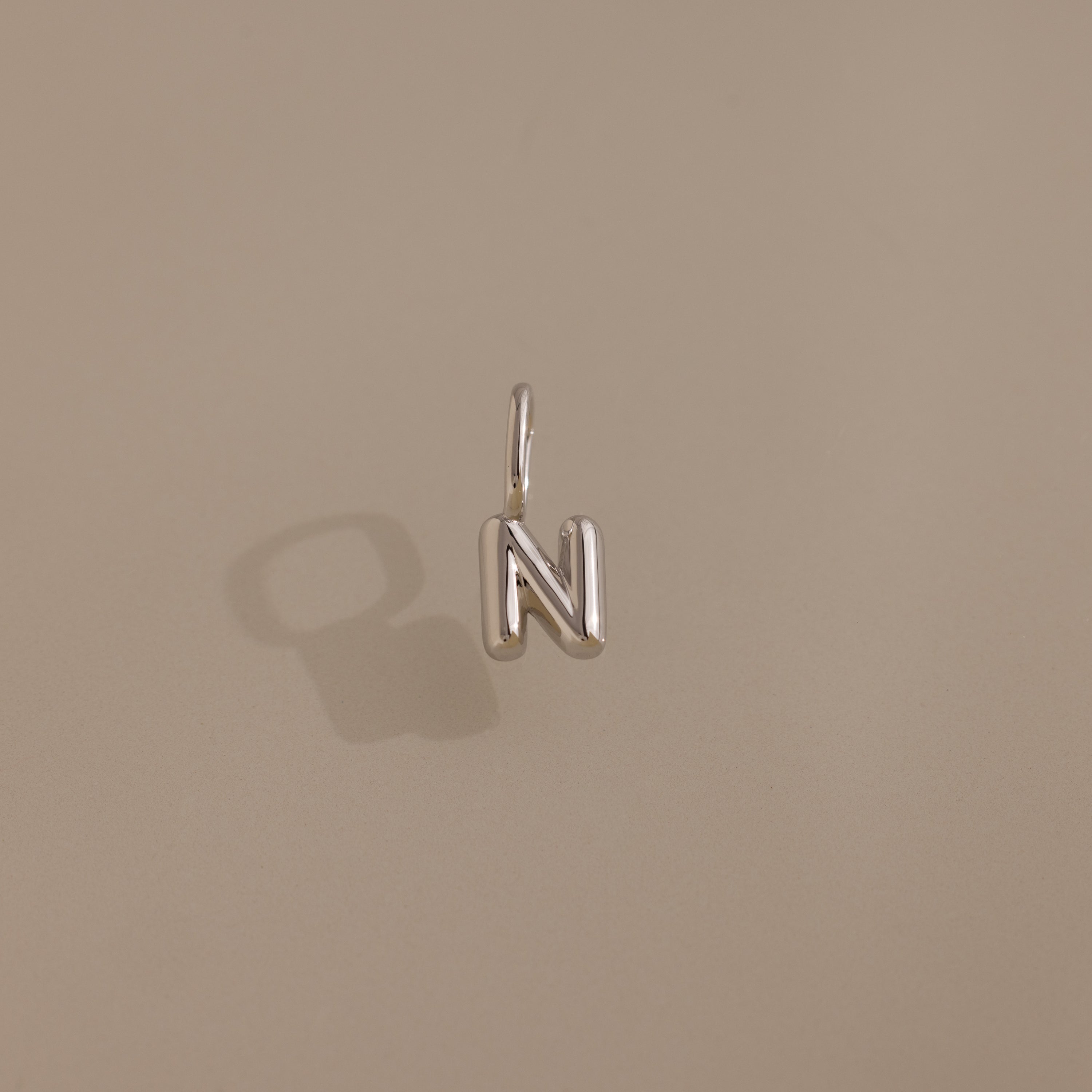 The Bubble Letter Charm in Sterling Silver, shaped as the letter N, rests on a neutral beige background—ideal for fans of personalized jewelry.