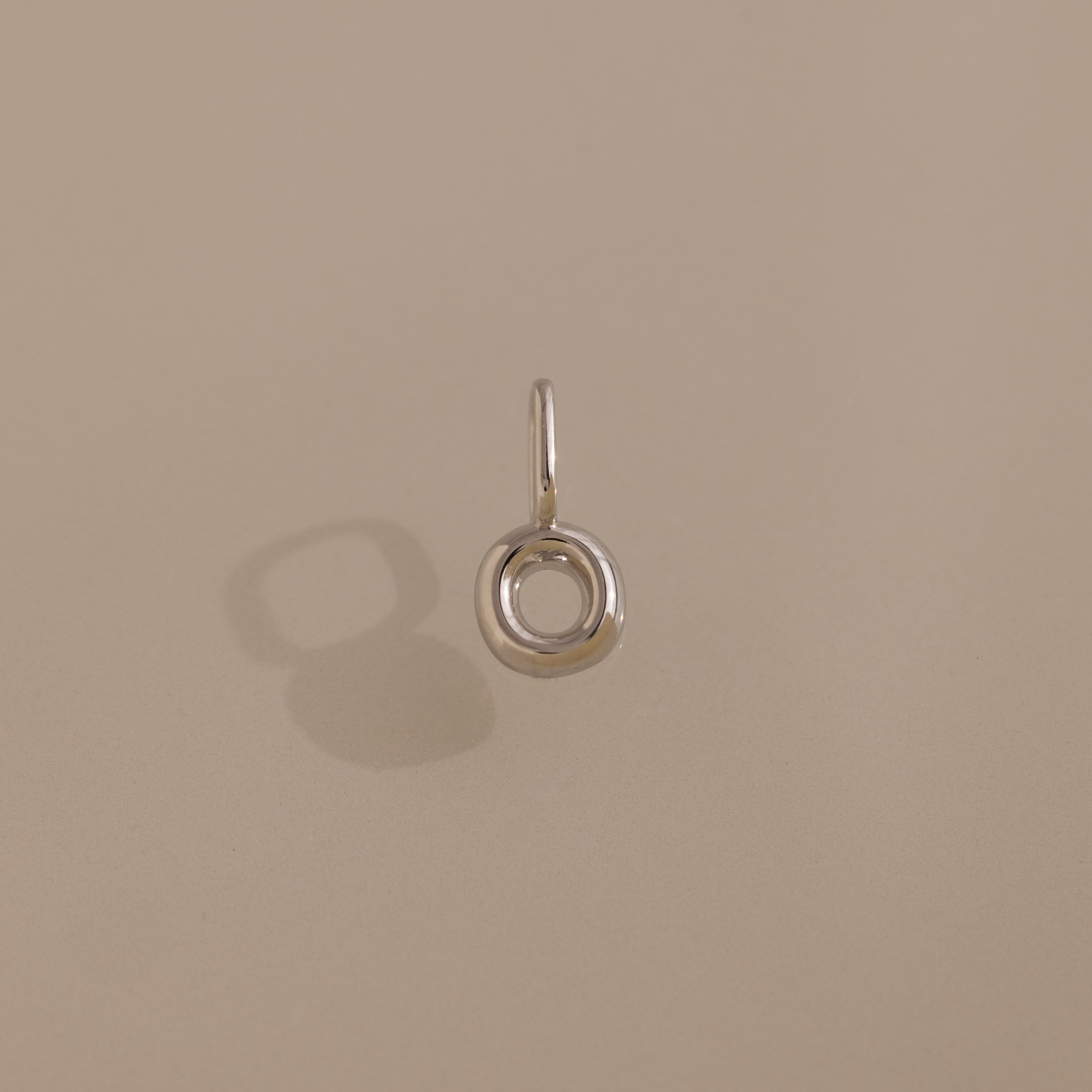 The Bubble Letter Charm in Sterling Silver, a small circular pendant, casts a shadow on a beige surface.