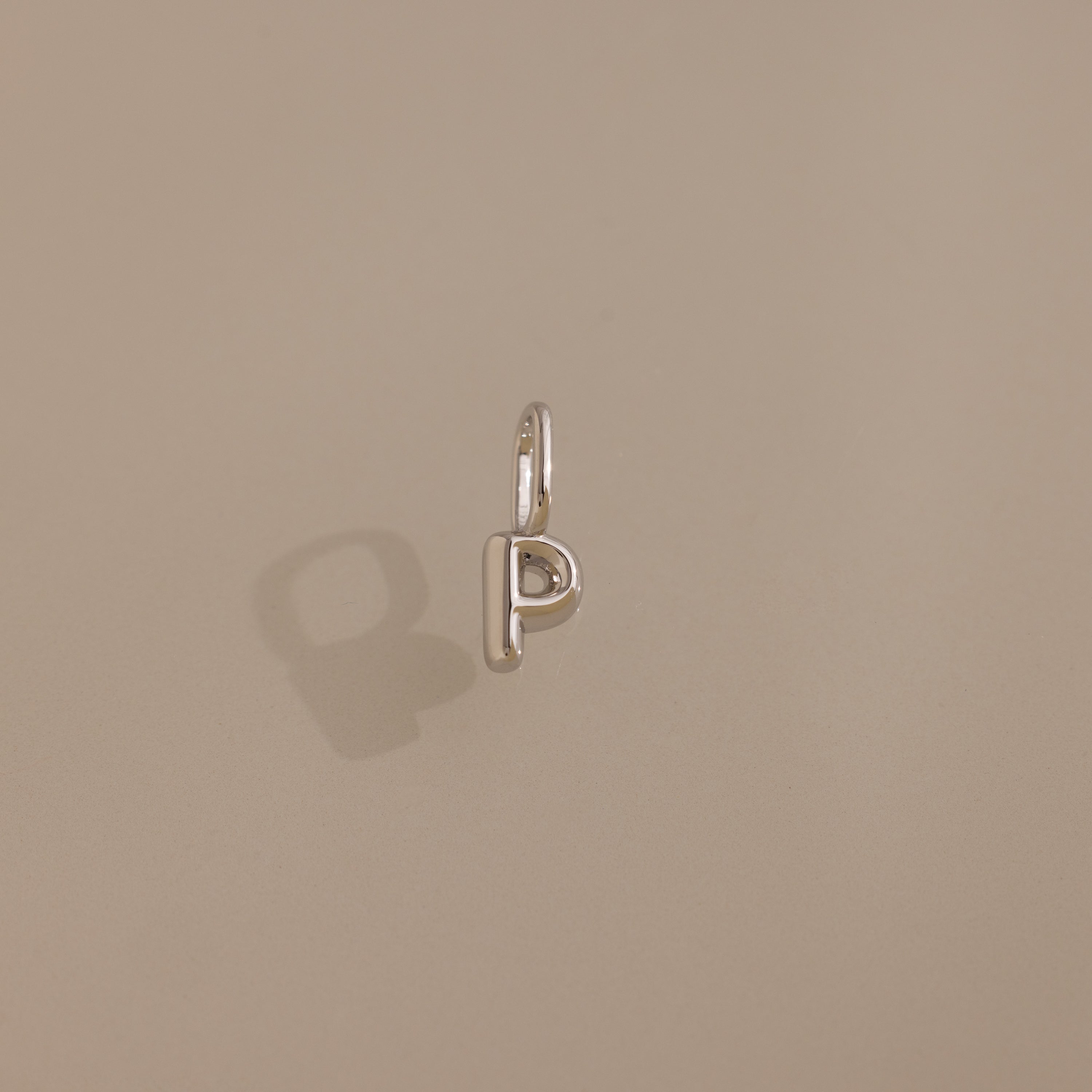 The Bubble Letter Charm in Sterling Silver casts a shadow on a plain beige surface, making it ideal for anyone seeking personalized jewelry.
