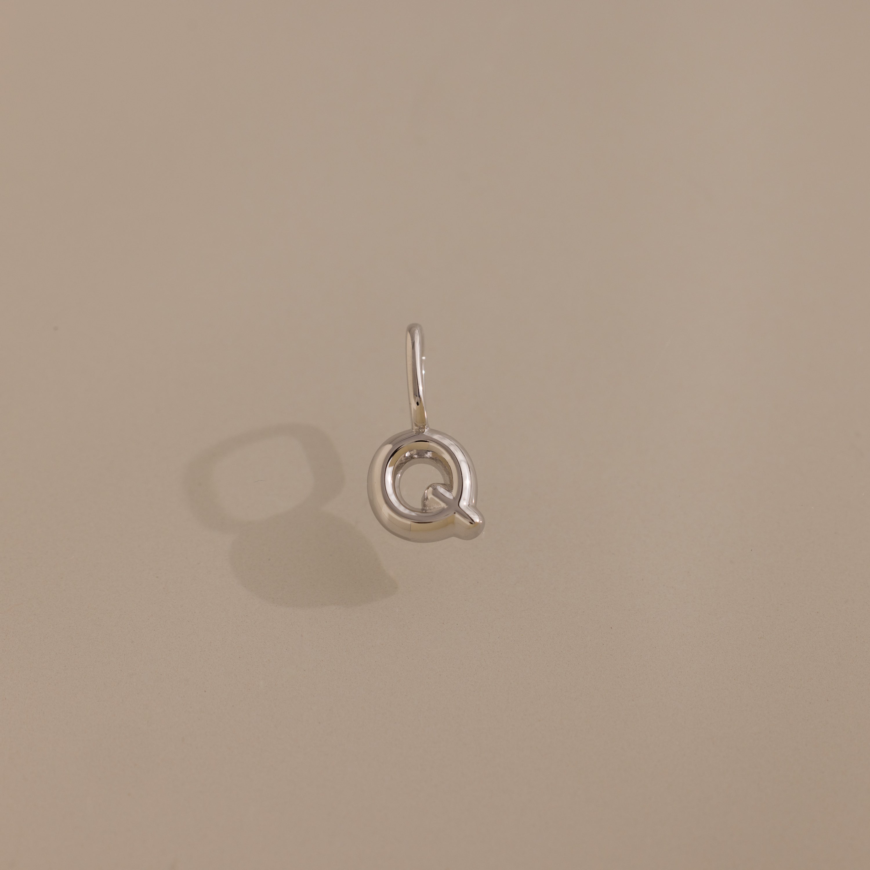 The Bubble Letter Charm in Sterling Silver, shaped like the letter Q, sits on a neutral beige background—ideal for anyone looking for playful, personalized jewelry.