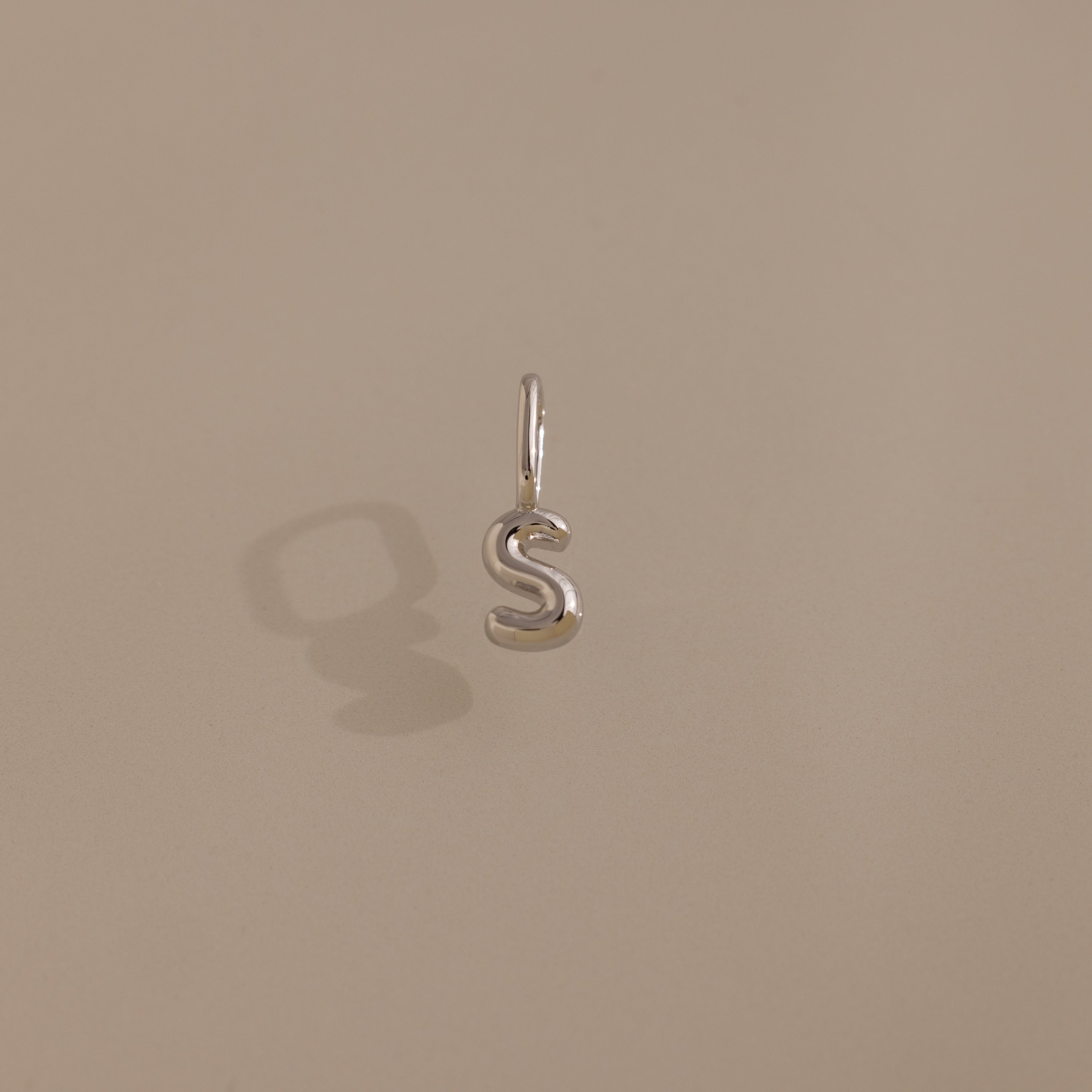 The Bubble Letter Charm in Sterling Silver, shaped like an S, rests on a neutral beige background with a soft shadow—ideal for lovers of personalized jewelry.