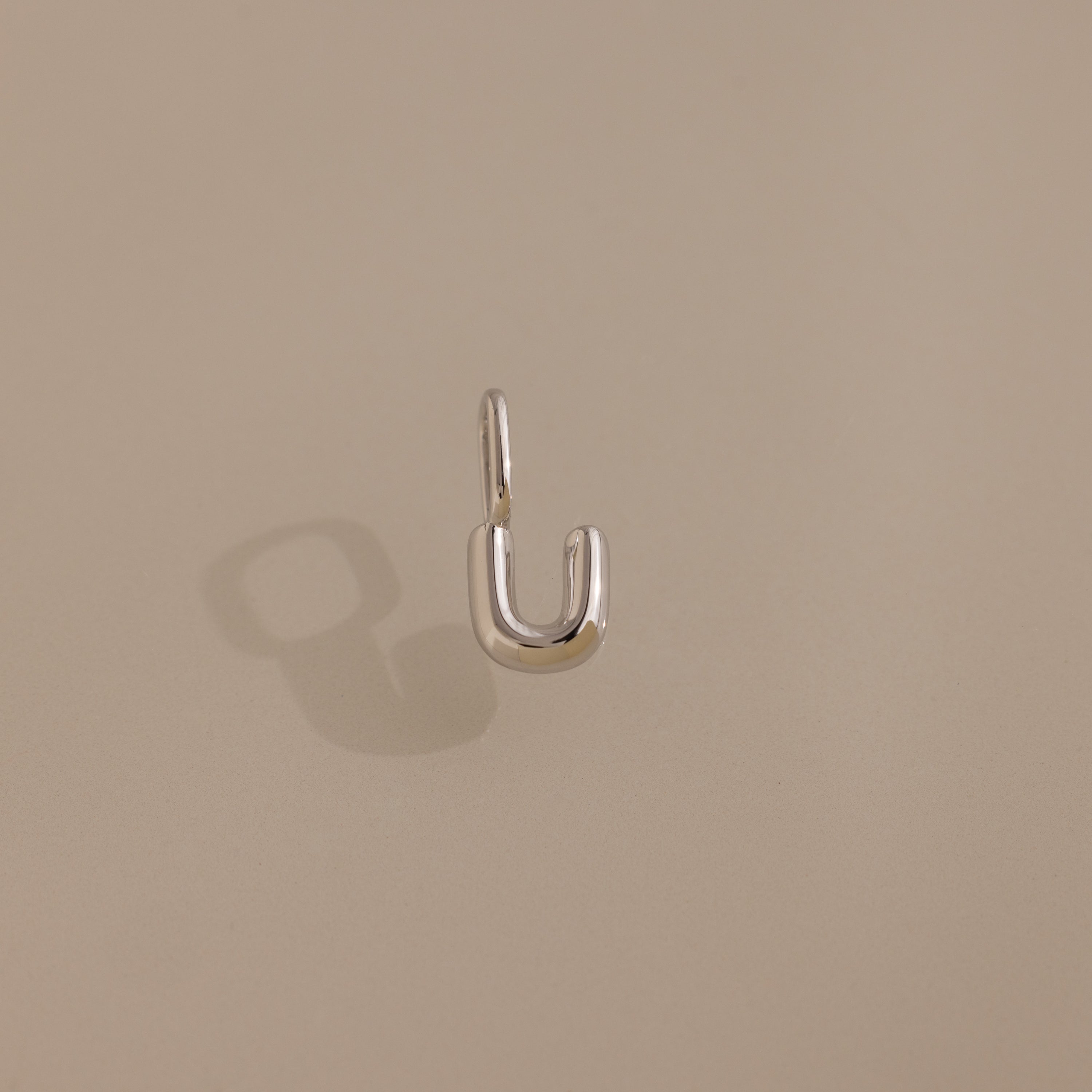 The Bubble Letter Charm in Sterling Silver, a small U-shaped earring with a subtle letter detail, rests on a beige background with its shadow softly beside it.