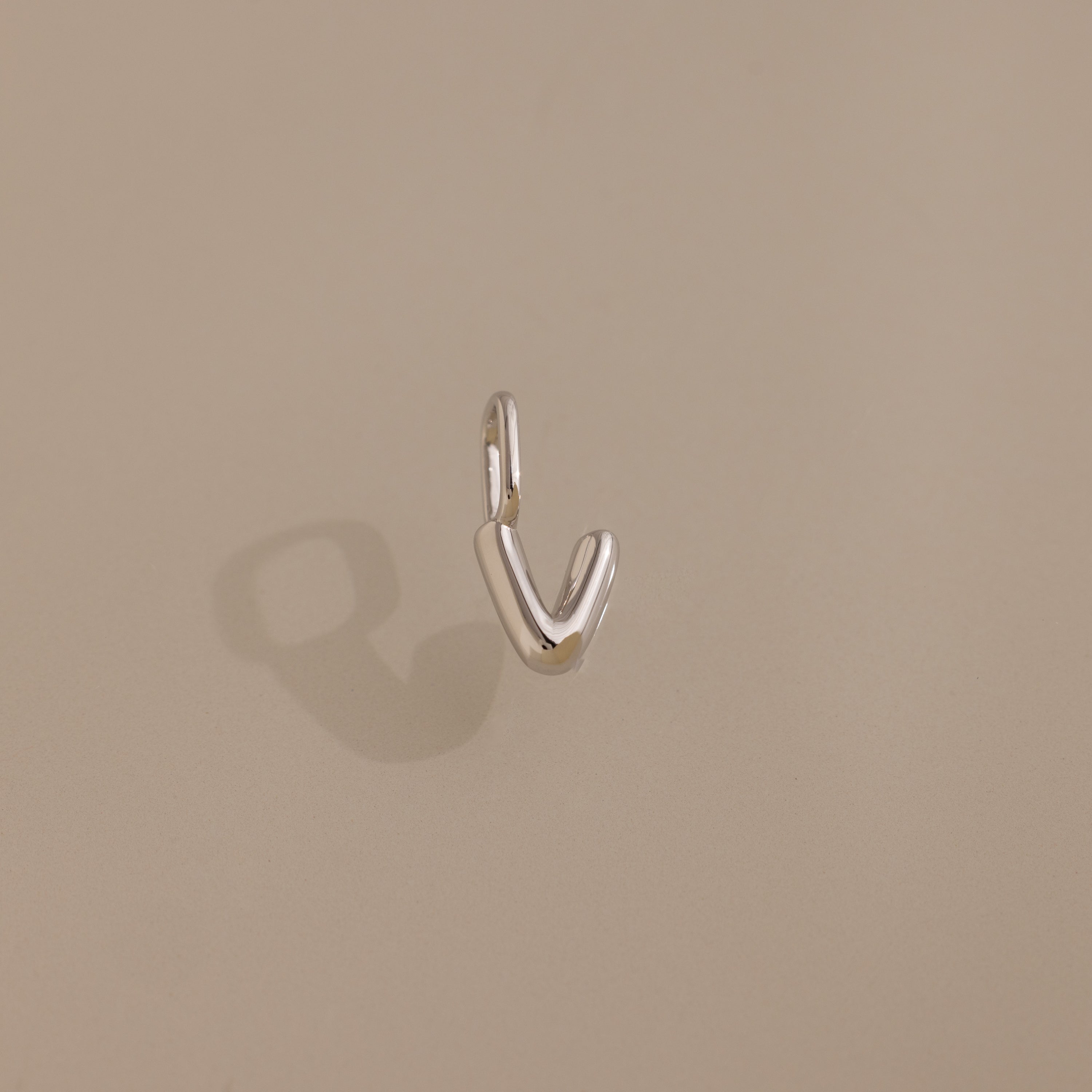 The Bubble Letter Charm in Sterling Silver, a small V-shaped earring with a delicate letter charm, casts a soft shadow on a beige surface.