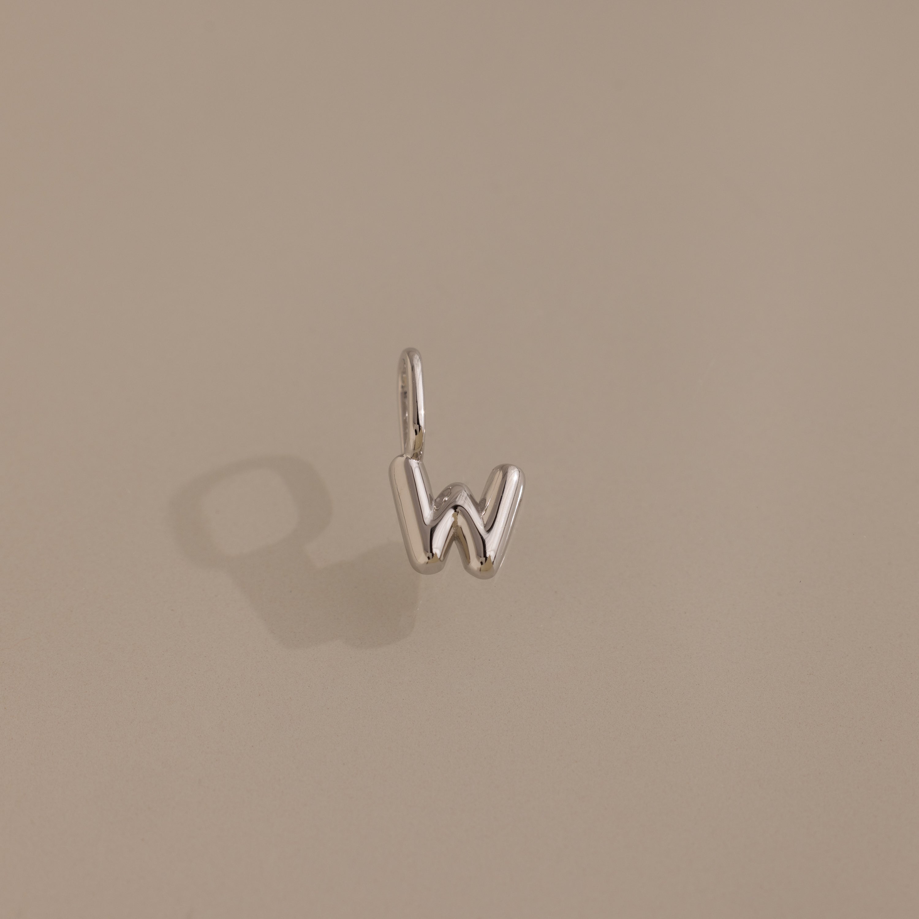 The Bubble Letter Charm in Sterling Silver, shaped like the letter W, is displayed on a plain beige background—ideal for adding a personalized touch to any jewelry collection.