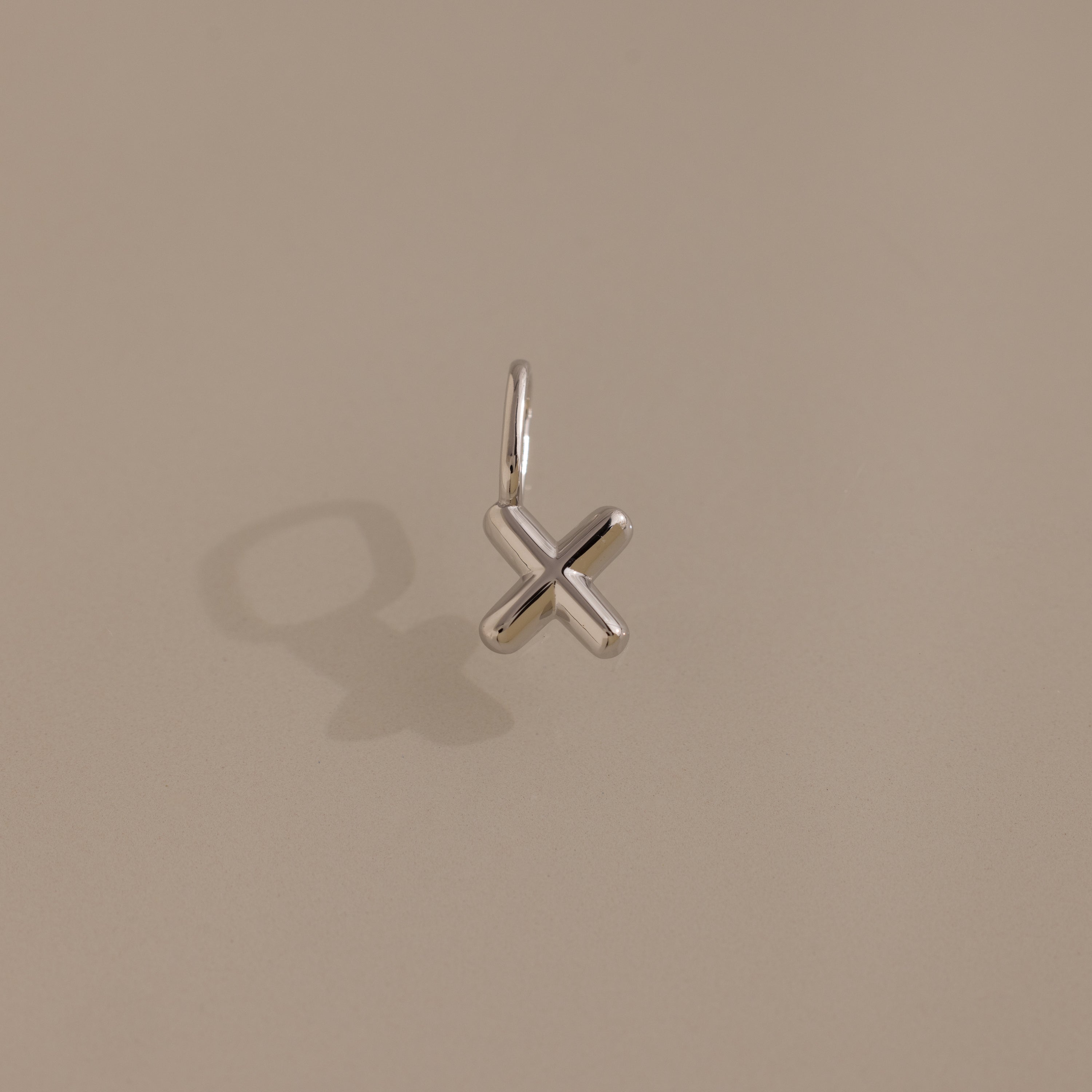 The Bubble Letter Charm in Sterling Silver features an elegant X-shaped design showcased on a light beige background.