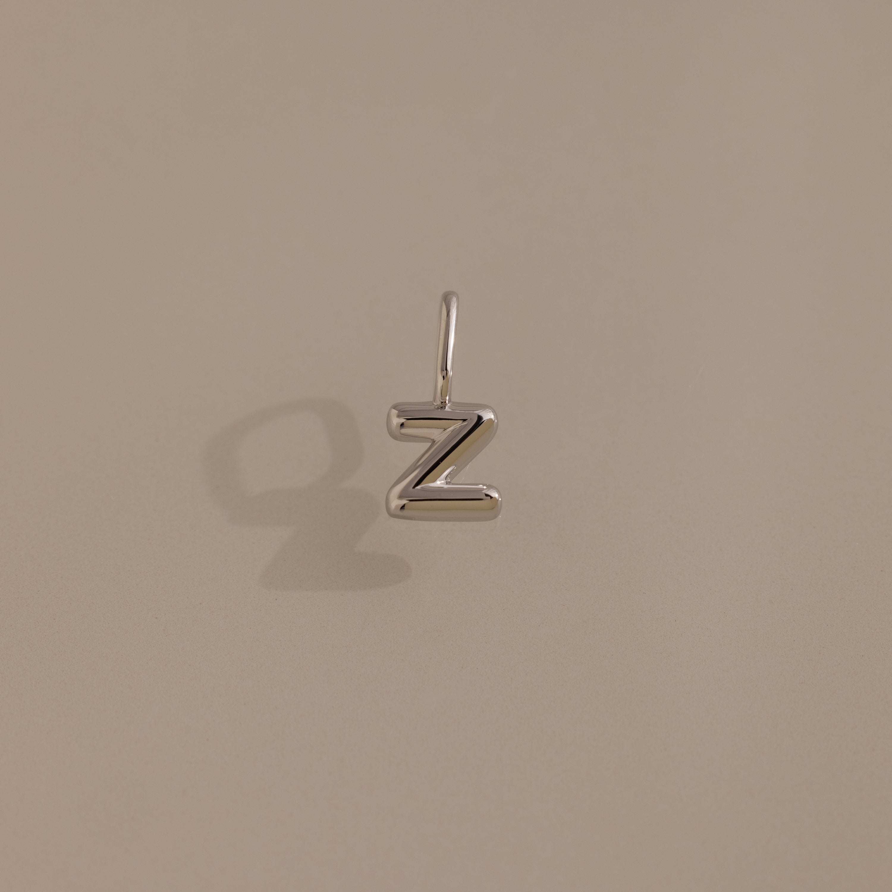 The Bubble Letter Charm in Sterling Silver, shaped like the letter Z, sits on a neutral beige background—ideal for anyone seeking personalized jewelry.