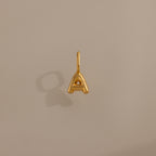 Bubble Letter Charm in 18K Gold shaped as an "A" shown on a plain beige background, casting a subtle shadow to the left.
