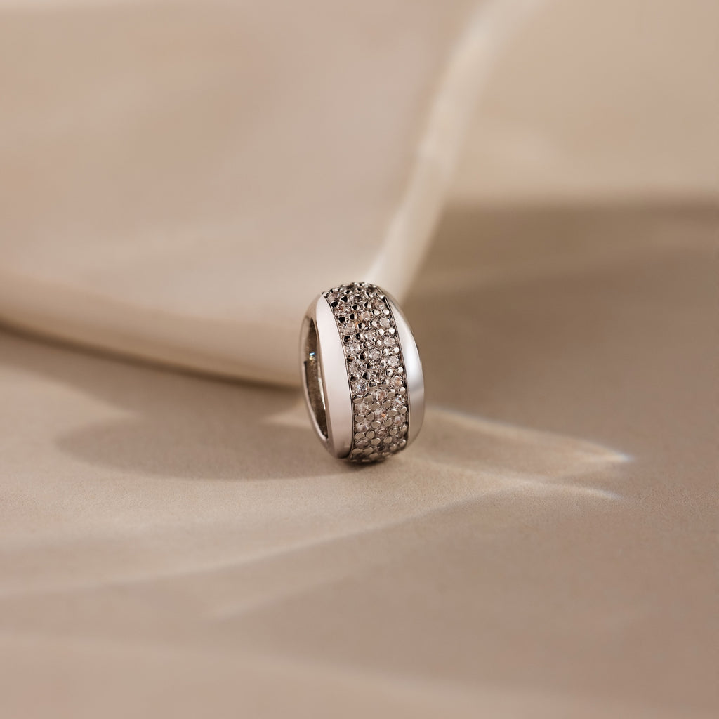 The Bold Pave Dome Charm in Silver features rows of sparkling small diamonds on a soft beige fabric background, adding timeless sophistication to any look.