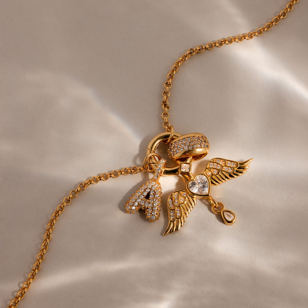 The Bold Pave Dome Charm in 18K Gold features a letter A, wing, heart, and ring—each adorned with sparkling stones. This personalized accessory shines with style and meaning.