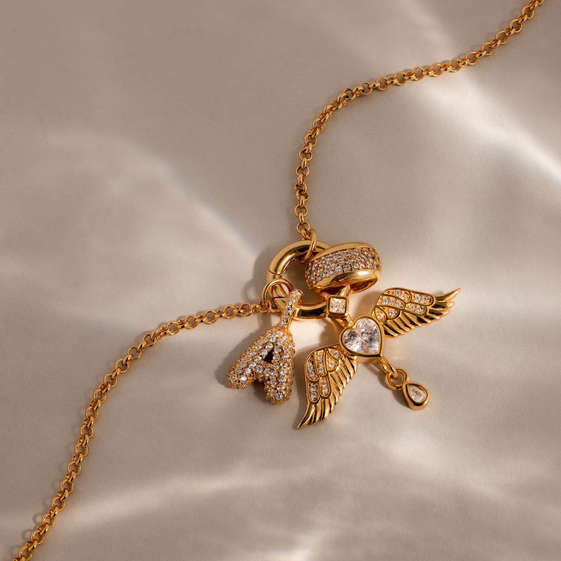The Bold Pave Dome Charm features a gold necklace with diamond-studded letter A, ring, and angel wings with heart charms—ideal for expressing love and style in one elegant piece.