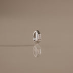 The Baguette Diamond Charm in Silver stands upright on a reflective beige surface, catching the light with its elegant rectangular gemstone and modern design.