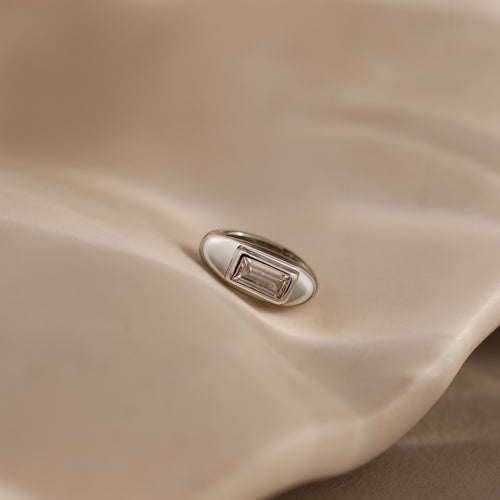 The Baguette Diamond Charm in Silver, featuring a rectangular clear stone, rests elegantly on a smooth beige surface, making it a timeless jewelry piece.