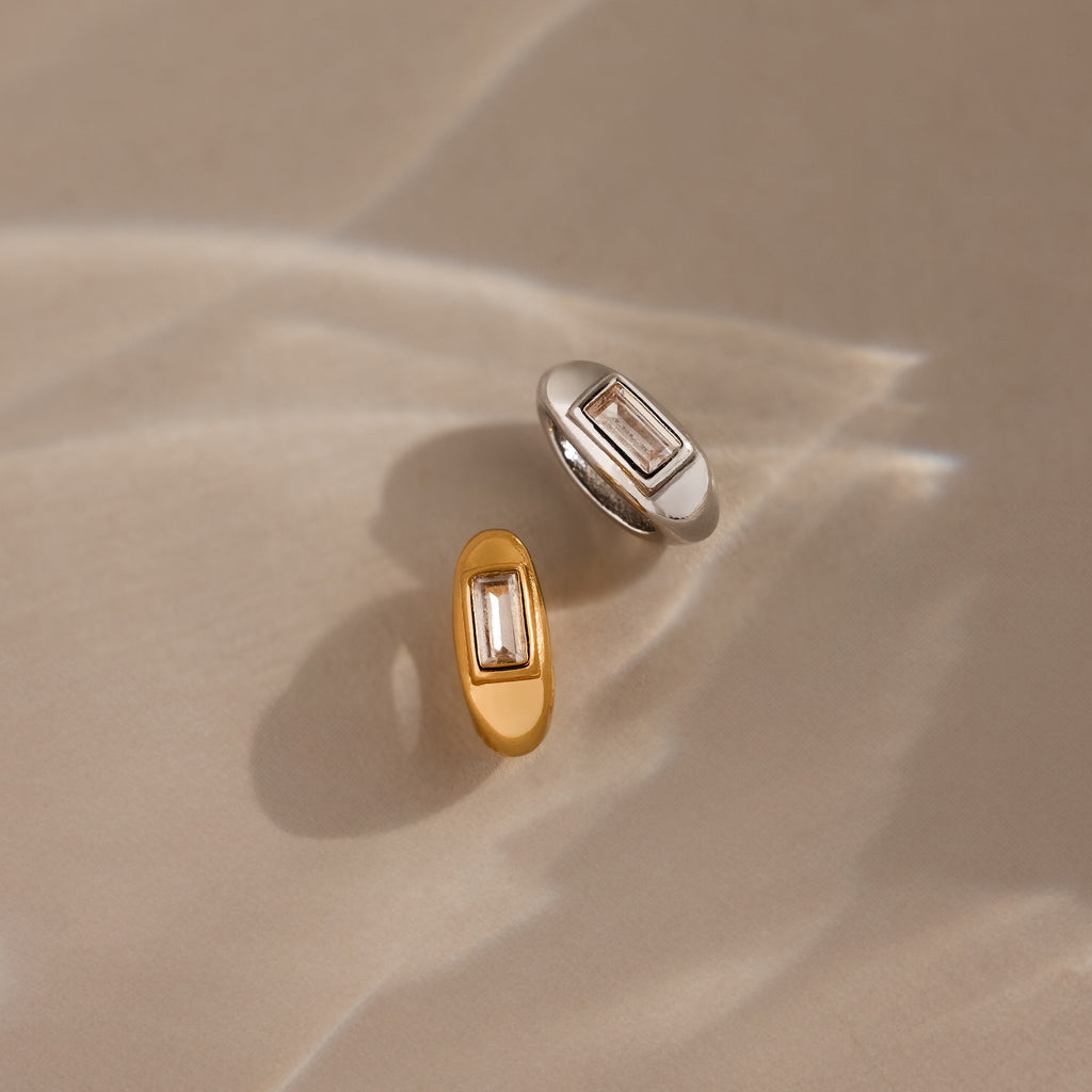 A gold and a silver oval ring, each featuring a rectangular clear stone resembling the Baguette Diamond Charm, are displayed on a neutral surface. No additional product description is available.