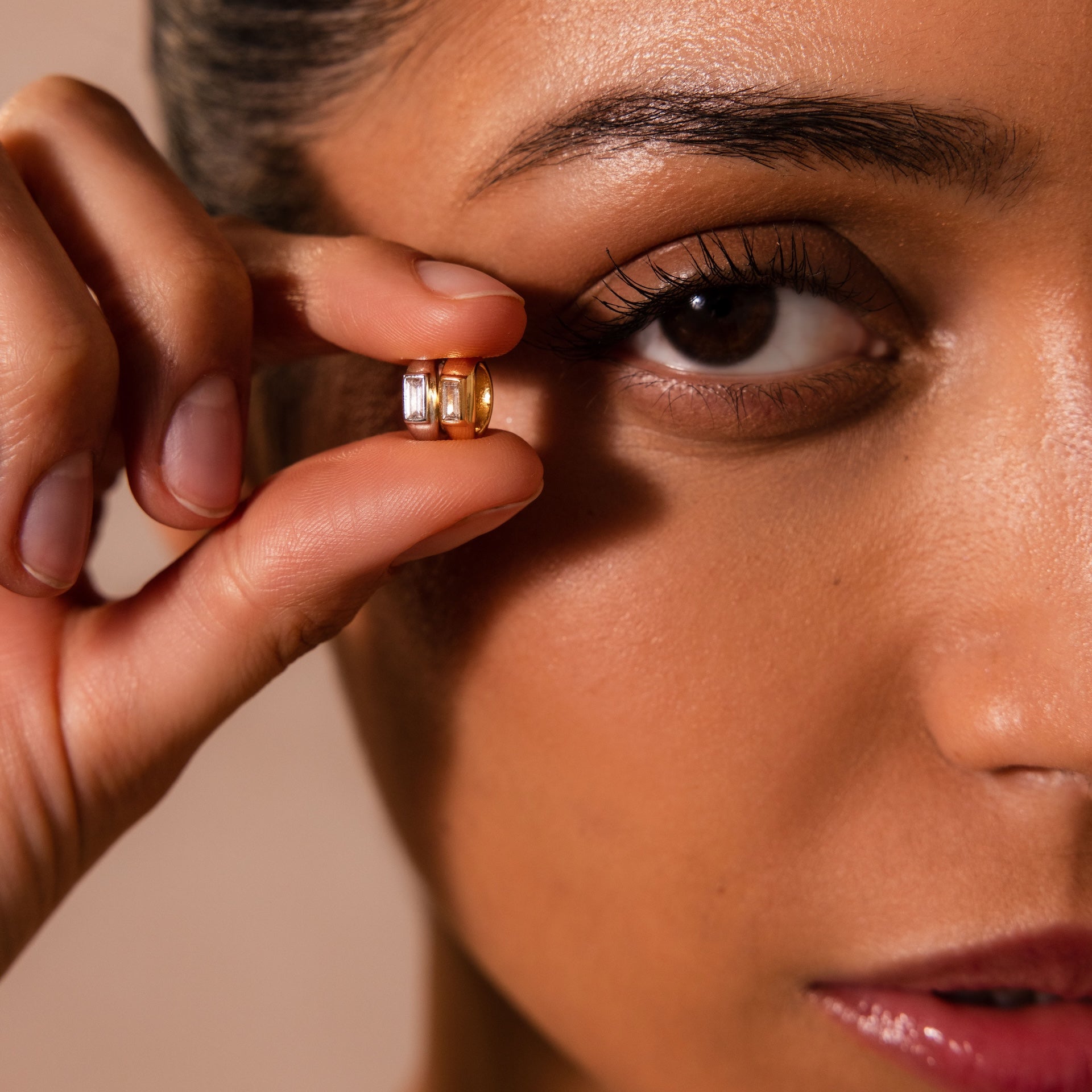 A woman holds two small Baguette Diamond Charms near her eye in a close-up shot, highlighting the sparkling details of the jewelry.