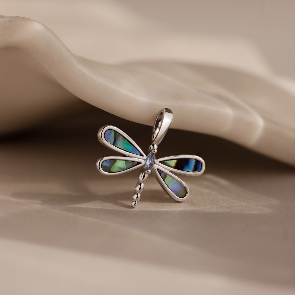 The Abalone Dragonfly Charm features shimmering, iridescent wings and rests elegantly on a smooth beige surface, reflecting nature-inspired beauty.
