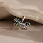 The Abalone Dragonfly Charm features shimmering, iridescent wings and rests elegantly on a smooth beige surface, reflecting nature-inspired beauty.